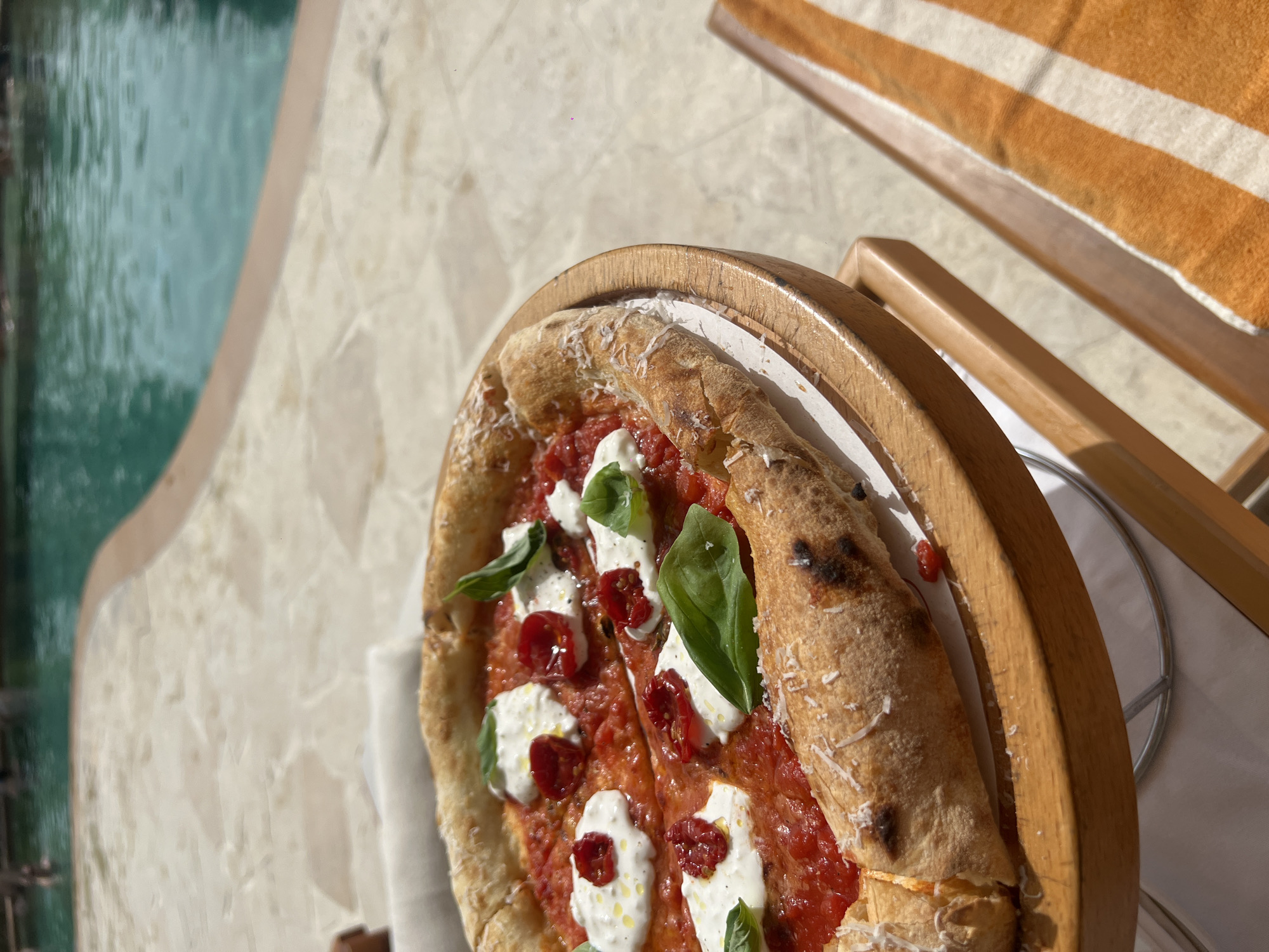 The image shows a pizza on a wooden tray, placed on a beach chair. The pizza appears to be topped with tomato sauce, mozzarella, sun-dried tomatoes, and basil. The chair is next to a swimming pool with turquoise water. The location is possibly in a resort in Dubai, United Arab Emirates, as suggested by the presence of the pool, beach chair, and the style of the pizza.