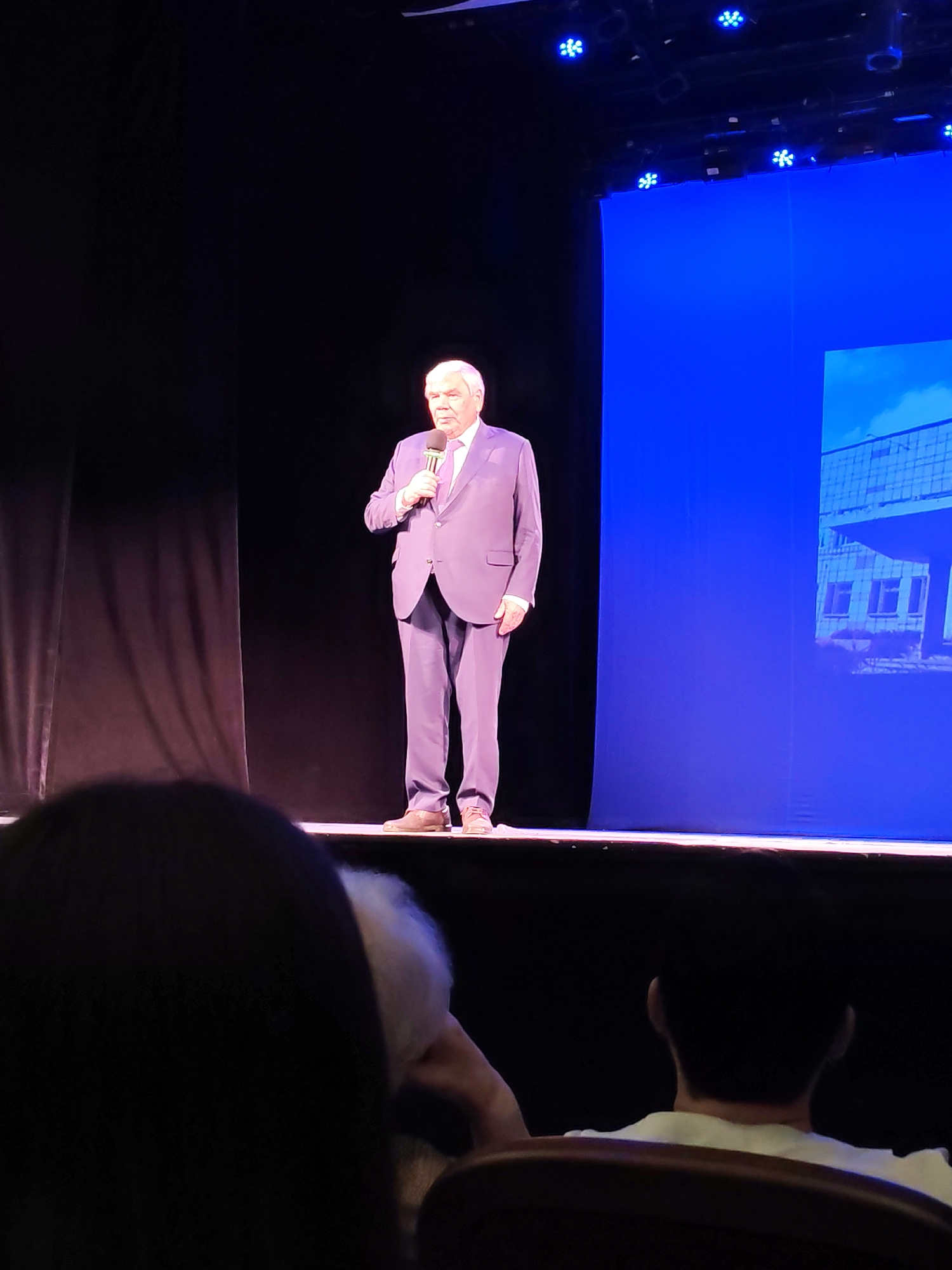 An older man in a light purple suit and tie is standing on a stage in front of a dark curtain and a bright blue backdrop. He is holding a microphone in his right hand and appears to be speaking. The stage is lit by spotlights from above. The image was taken at the Chaikovsky Concert Hall in Chaikovsky, Russia. In the foreground, the backs of several audience members are visible, with one person in the lower right appearing to be watching the speaker.