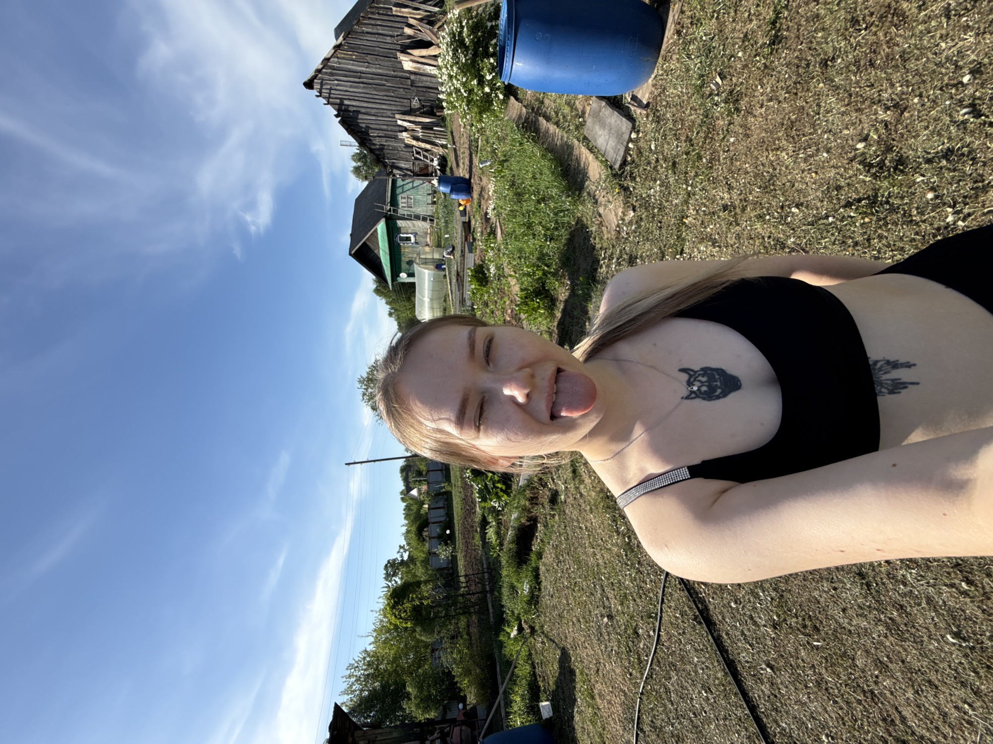 A medium shot shows a woman with blonde hair and fair skin in an outdoor setting. She is wearing a black sports bra and has tattoos on her upper chest and arm. She is sticking her tongue out and winking.
The background includes a rural landscape with wooden buildings and green vegetation. A blue barrel is visible in the foreground. A blue sky with sparse clouds occupies the upper portion of the frame. The location is in the city of Unknown, Russia.