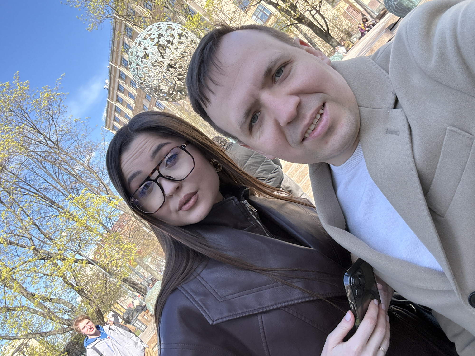 A selfie of a man and a woman with a distinctive building and sculpture in the background, taken at an oblique angle under daylight conditions. The man, on the right, wears a light brown coat over a white shirt and smiles, while the woman on the left wears glasses and a brown leather jacket. The woman holds a dark-colored smartphone. Behind them, a tall building with numerous windows rises into a bright blue sky. A large, spherical metal sculpture is positioned near the top of the building and to the left of the pair. Bare trees with nascent leaves are visible in the foreground and around the building. In the background, additional people are present, some seated on a ledge. A male in a gray jacket is seen in the left lower portion of the image.