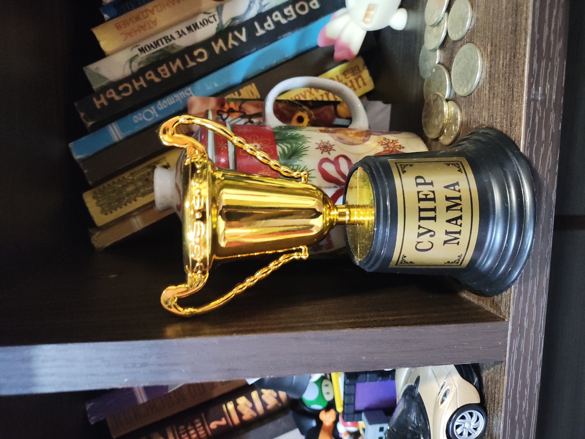 The image shows a shelf in a home, filled with various items. A golden trophy with "SUPER MAMA" inscribed on a black base is prominently displayed. Behind the trophy is a mug with a festive design, possibly Christmas-themed. Several books are stacked on the shelf, with titles in a language other than English. A small plush toy is visible near the top. A collection of coins is scattered beside the trophy. On the lower shelf, there are more books and toys, including a toy car. The location appears to be a residential interior.