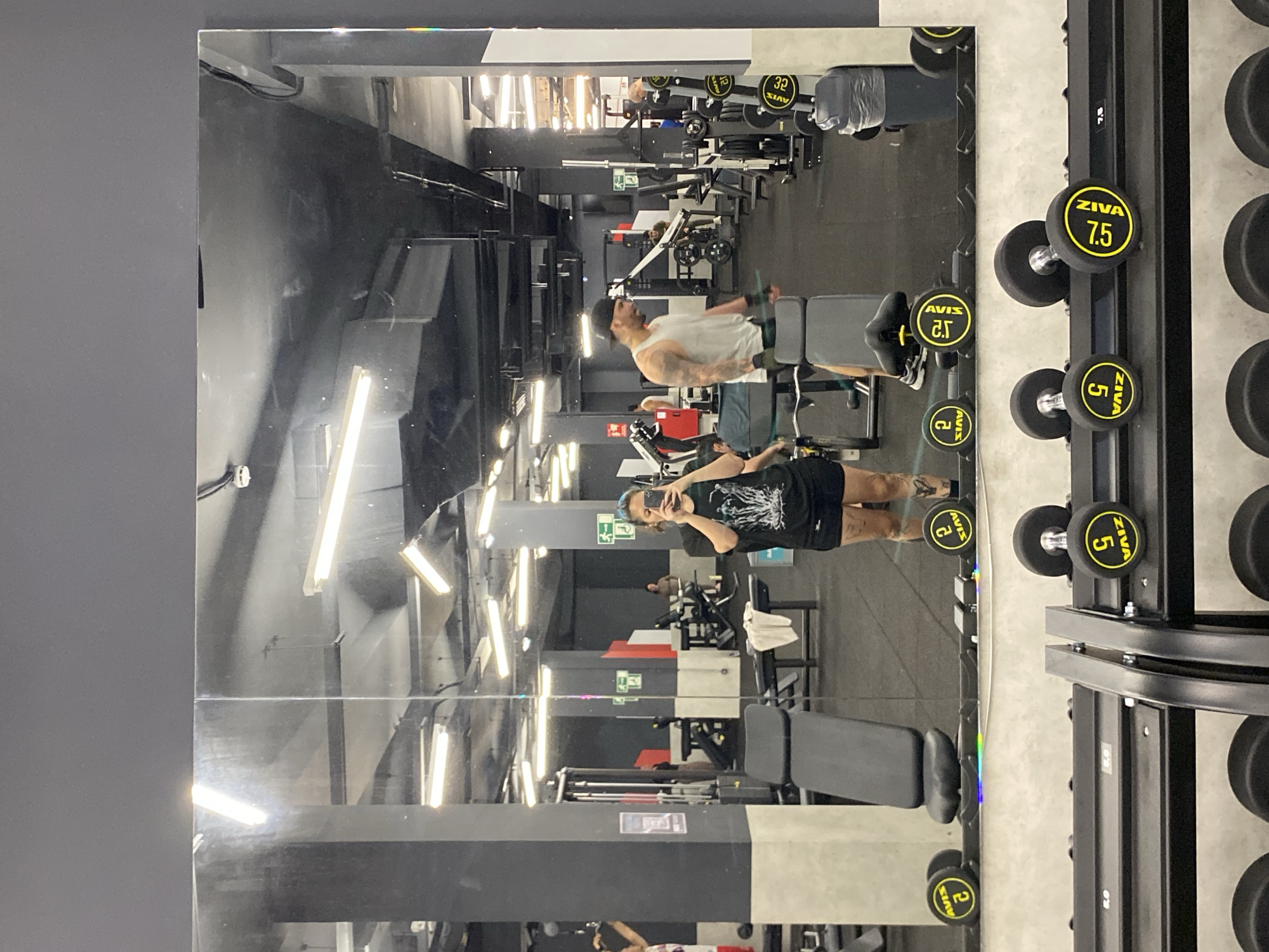 This media file captures a mirror selfie taken inside a gym located in Gijón/xixón, Spain. The image is oriented sideways, but depicts a large reflective mirror on the left, with a gray wall to its left.
The primary subject is a person taking the selfie, reflected in the mirror. They are wearing a black t-shirt with a white design and black shorts, holding a smartphone up to capture their reflection. Their face is mostly obscured by the phone. In the background of the reflection, another person, a man wearing a white tank top and a black baseball cap, is visible, possibly exercising or resting near gym equipment.
The scene is a modern gym with dark walls, a gray floor, and bright fluorescent strip lighting on the ceiling, which reflects strongly in the mirror. Various weight machines and benches are visible in the reflection. To the right of the frame, a black dumbbell rack holds several dumbbells.
Visible text on the dumbbells includes "ZIVA" and "AVIS", indicating the brand, along with weight markings such as "7.5", "5", "2.5", and "2". The number "32" is also visible on some equipment in the deeper background. The activity is primarily the act of taking a mirror selfie within a fitness environment.