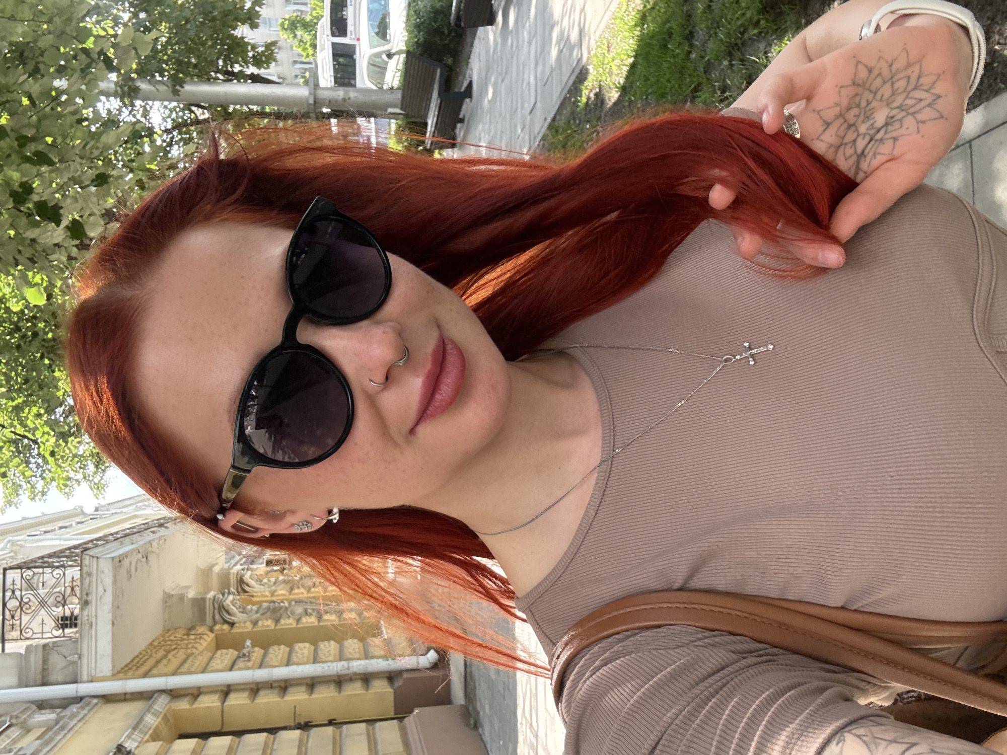 A young woman with auburn hair, sunglasses, and a cross necklace is taking a selfie. Her hand, adorned with a floral mandala tattoo and a ring, is holding her hair. The scene is set outdoors. The woman is wearing a taupe shirt and a brown leather bag strap is visible over her shoulder. In the background, there are buildings with ornate architectural details, trees, benches, a sidewalk, and a white van. The location appears to be urban. Based on image analysis, it is difficult to confidently pinpoint the location as Rostov-on-Don, Russia.