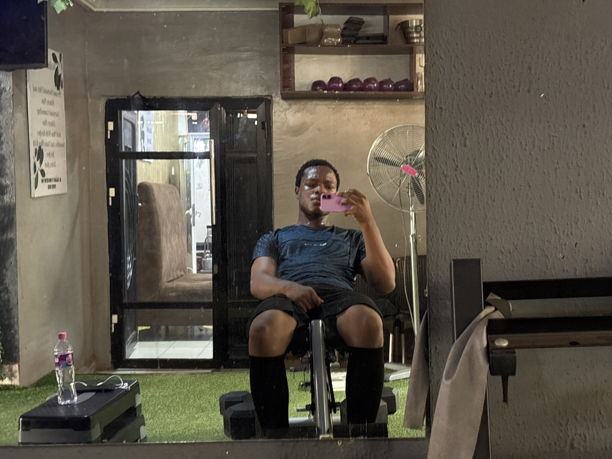 The image depicts a man taking a selfie in what appears to be a gym or fitness studio located in Enugu, Nigeria. The man is seated on a weight bench, holding a pink smartphone to take the picture. He is wearing a blue t-shirt, black shorts, and black compression socks. In the background, a large industrial fan is visible, along with shelves containing various items, including what seem to be purple exercise balls. To the left, a glass door leads to another area where a large, upholstered chair is visible. A water bottle sits on a black step platform in the foreground. The environment has a textured, grey wall and artificial turf flooring. The reflection in the mirror suggests the gym is indoors, with artificial lighting.