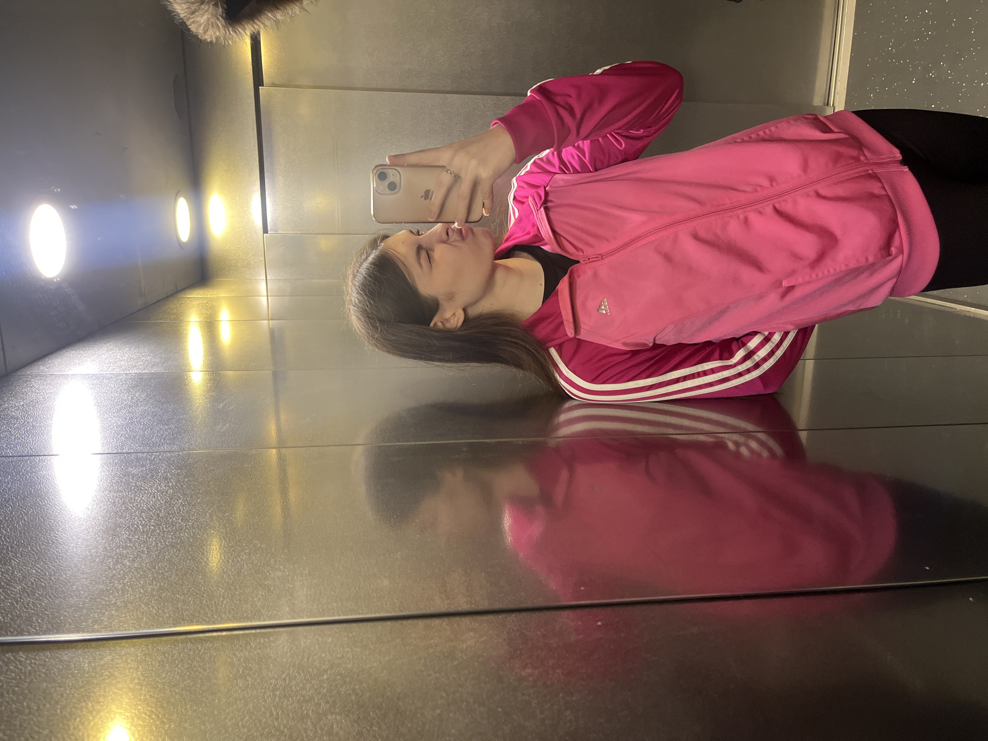 A young woman is taking a selfie in what appears to be an elevator. She is wearing a pink Adidas tracksuit and black leggings. She has brown hair and is pouting at the camera. The elevator has stainless steel walls and a mirrored finish on the floor. There are several circular lights on the wall behind her. The location of the image in the city of Saint Petersburg, Russia cannot be determined with certainty.