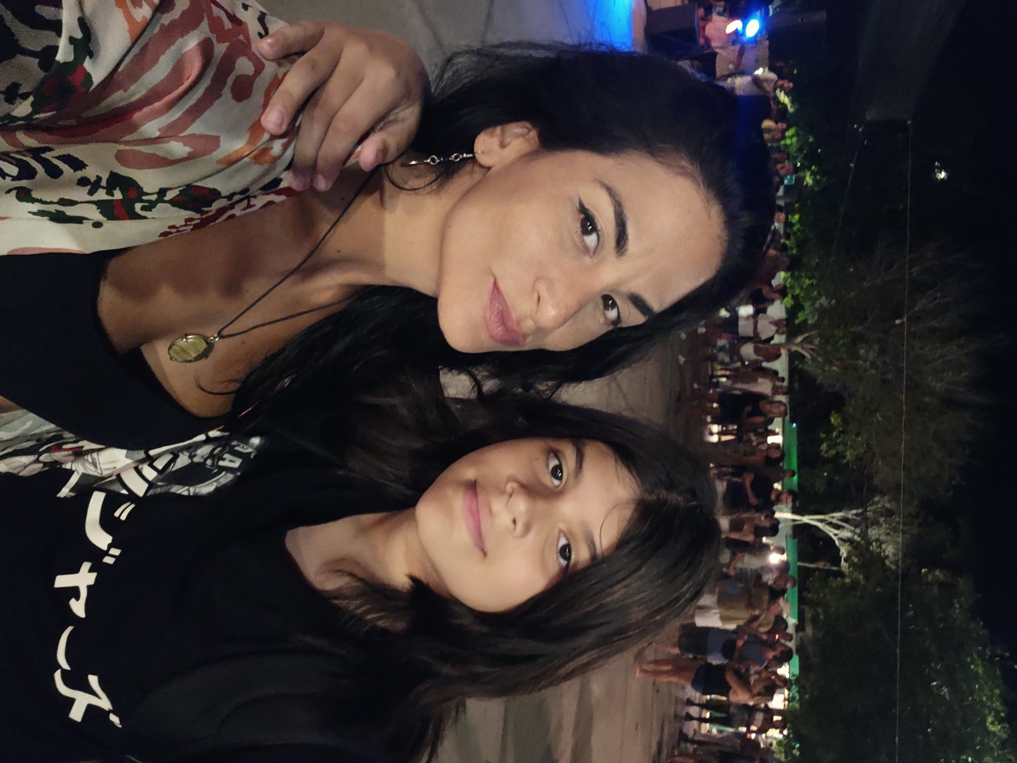 The image displays two individuals in the foreground, an adult female and a younger individual, positioned closely together in what appears to be an outdoor nighttime event. The adult female, located on the left, has long dark hair, defined eyeliner, and wears a light-colored, multi-patterned kimono over a black top, accessorized with a black necklace featuring an oval pendant and a silver dangle earring. She is looking towards the camera with a slight smile and has her right arm extended around the younger individual's left shoulder. The younger individual, on the right, also possesses long dark hair and wears a black t-shirt featuring white Japanese characters across the chest, along with a black backpack. This individual maintains a neutral expression while looking towards the camera.
The background is dark and blurred, populated by a crowd of numerous individuals standing and mingling, suggesting a public gathering or festival. Ambient artificial lighting illuminates parts of the scene, with a prominent blue light source visible on the far left. Dark trees are visible against the night sky in the mid-ground. The ground surface appears to be unpaved. The specific location within the city of Unknown, Unknown, is not identifiable; however, the scene characteristics are consistent with an outdoor night market, concert, or festival environment.