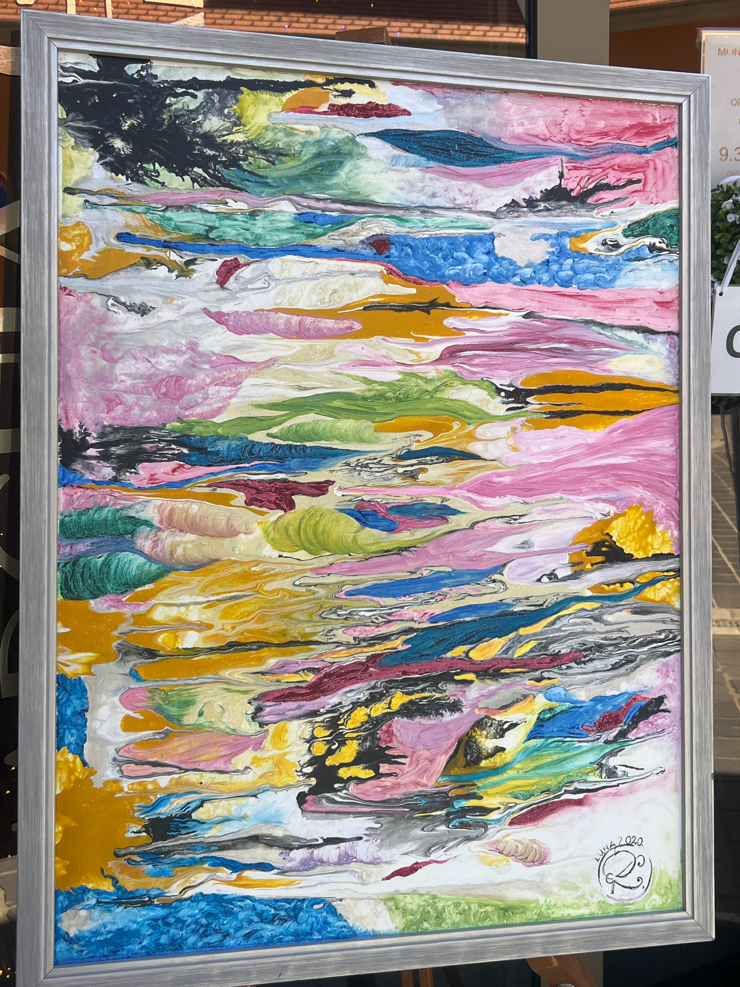 The image is a vertical, medium shot of an abstract painting framed in silver and displayed outdoors, likely in a city setting. The painting itself is a vibrant and chaotic composition of swirling, layered colors, predominantly featuring shades of pink, blue, green, yellow, and black. The paint appears thick and textured, with a sense of movement and fluidity. A signature and date, "LUHA 2020" along with a circular emblem, are visible in the bottom right corner of the painting.
In the background, a brick building with a tiled roof is visible at the top. To the right of the painting, a white sign with black text is partially visible, with the number "9.3" clearly readable. The sign appears to be an advertisement or informational display. There are also hints of foliage and possibly a white hanging flower arrangement to the right of the painting. The overall impression is that the painting is being exhibited or sold outdoors, possibly in a market or along a street.
Without additional contextual information or specific landmarks, the exact location in the city of Budapest, Hungary, cannot be determined from the image alone. The scene depicts a visual art display rather than a specific identifiable landmark within Budapest.