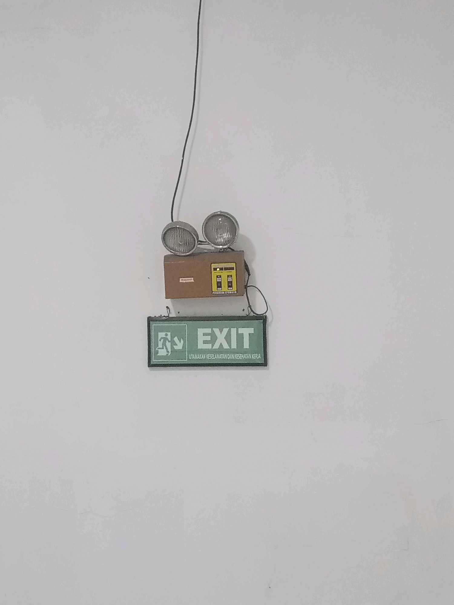 This image depicts an emergency exit sign mounted on a white wall. Above the sign is an emergency light fixture with two adjustable spotlights. The exit sign itself is rectangular with a green border and a white interior. It features a pictogram of a person walking towards an arrow pointing downwards and the word "EXIT" in large white letters. Below "EXIT," there is smaller text in Indonesian that reads "UTAMAKAN KESELAMATAN DAN KESEHATAN KERJA," which translates to "PRIORITIZE SAFETY AND OCCUPATIONAL HEALTH." The overall scene appears to be indoors, possibly in a public building or workplace, given the nature of the signage. The lighting suggests it is daytime, but the interior environment is not clearly visible. The location is specified as Balaraja, Indonesia. There are no people or interactions visible in the image.