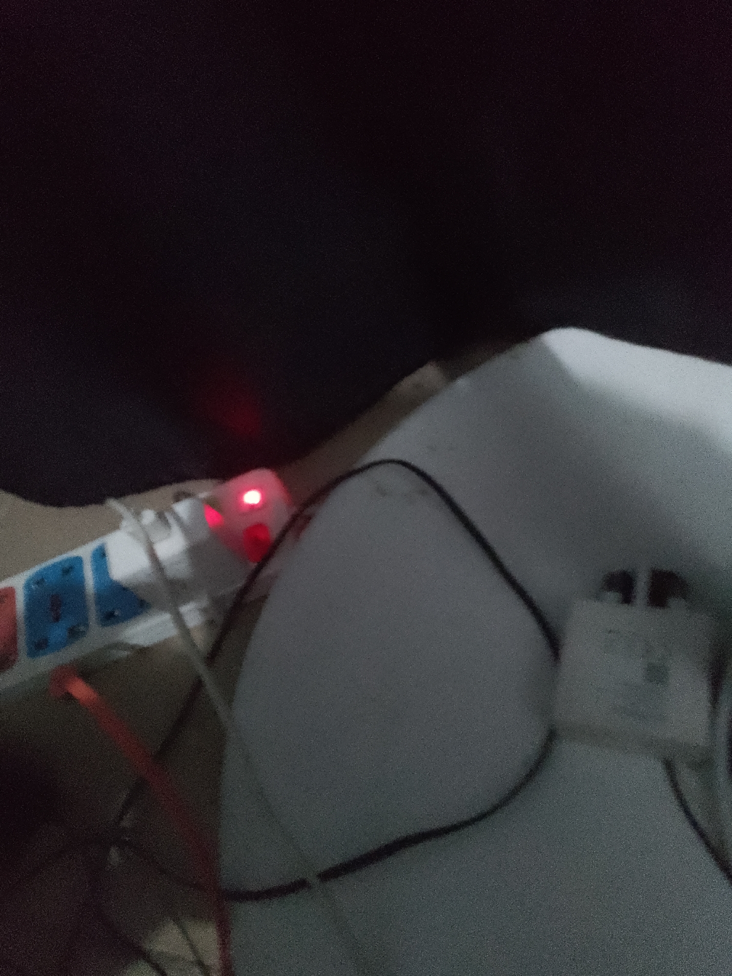 In the city of Jauro Yinu, Nigeria, a dimly lit scene captures a close-up view of electrical accessories. Dominating the frame is a white power strip with multiple outlets, some of which are occupied by electrical adapters. A small red indicator light glows on one of the adapters, suggesting it is active. Wires, including a red one, are connected to the power strip and trail away, disappearing into the shadows. The background is dark, with a soft, blurred surface that appears to be fabric, possibly a pillow or a blanket, on the right side of the image. The overall impression is one of a functional, yet somewhat cluttered, electrical setup in a low-light environment.