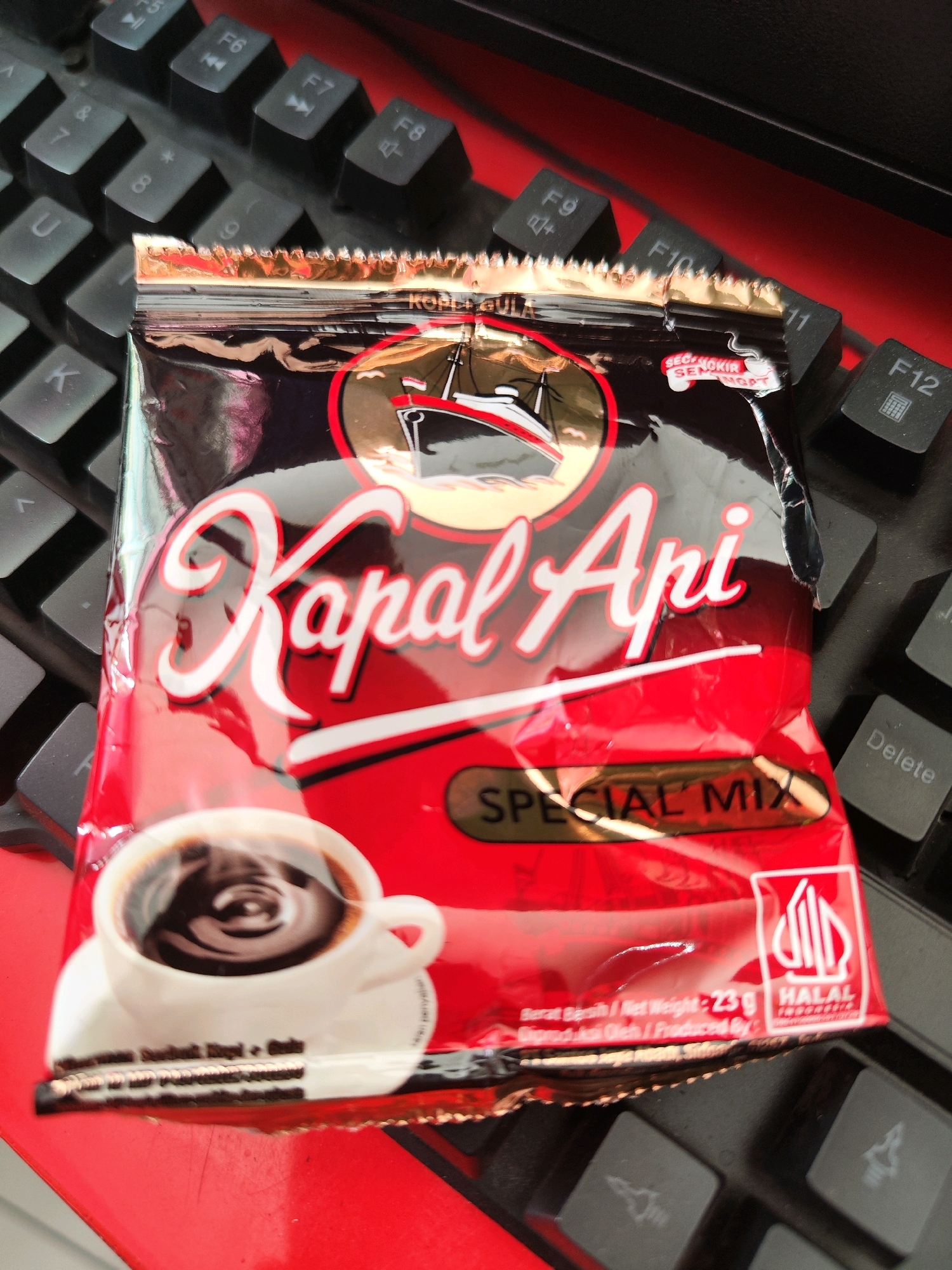 A close-up, overhead shot captures a packet of "Kapal Api Special Mix" coffee resting on a black keyboard with red accents. The packet is predominantly red with white lettering and a depiction of a steaming cup of coffee. The keyboard's keys are visible, including function keys F5 through F12, and other alphanumeric keys. The scene appears to be indoors, likely on a desk or table, with the lighting suggesting it could be in the city of Sragen, Indonesia, given the product packaging which is common in that region. There are no people or active interactions depicted in the image.