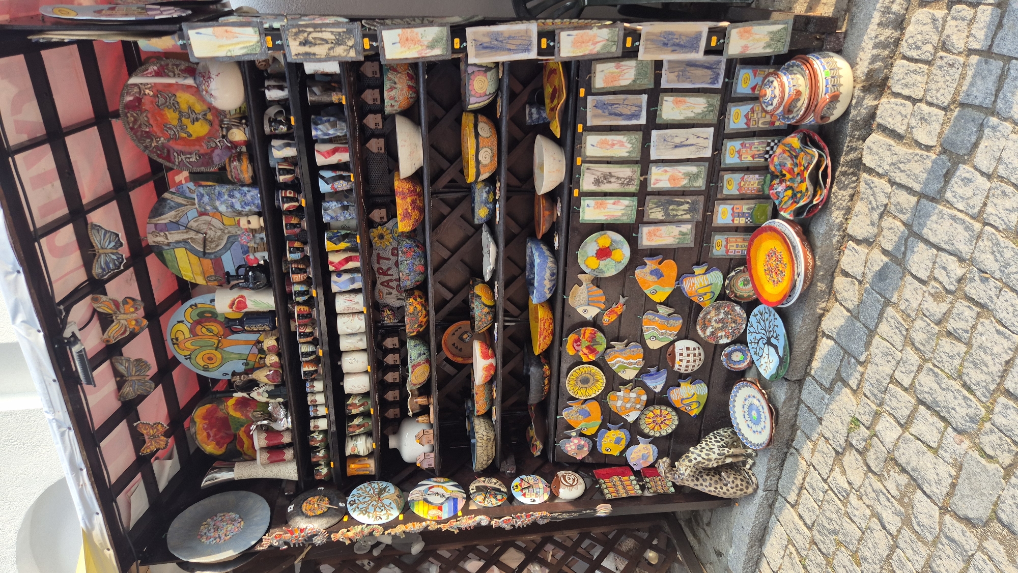 This is a medium shot, eye-level, outdoor photograph of a souvenir stall in Nesebar, Bulgaria. The stall is packed with various ceramic items, including plates, bowls, mugs, and decorative wall hangings. The items are displayed on wooden shelves and on the ground in front of the stall.
The stall is covered by a striped awning and decorated with several ceramic butterflies hanging from the ceiling. On the top shelf, there are several large, colorful ceramic plates and vases. Below these are rows of mugs and smaller decorative items. The lower shelves are filled with bowls and more plates, many of which are decorated with fish motifs.
In front of the stall, on a cobbled street, are more ceramic pieces, including several plates, a bowl, and a tiered, lidded pot. Two ceramic cheetahs are positioned on the left side of the stall, next to some hanging ornaments.
The lighting suggests it is daytime, with sunlight casting shadows. There are no people visible in the photograph. The background shows part of a building and a wooden lattice fence. The overall impression is of a vibrant and cluttered display of artisanal crafts.