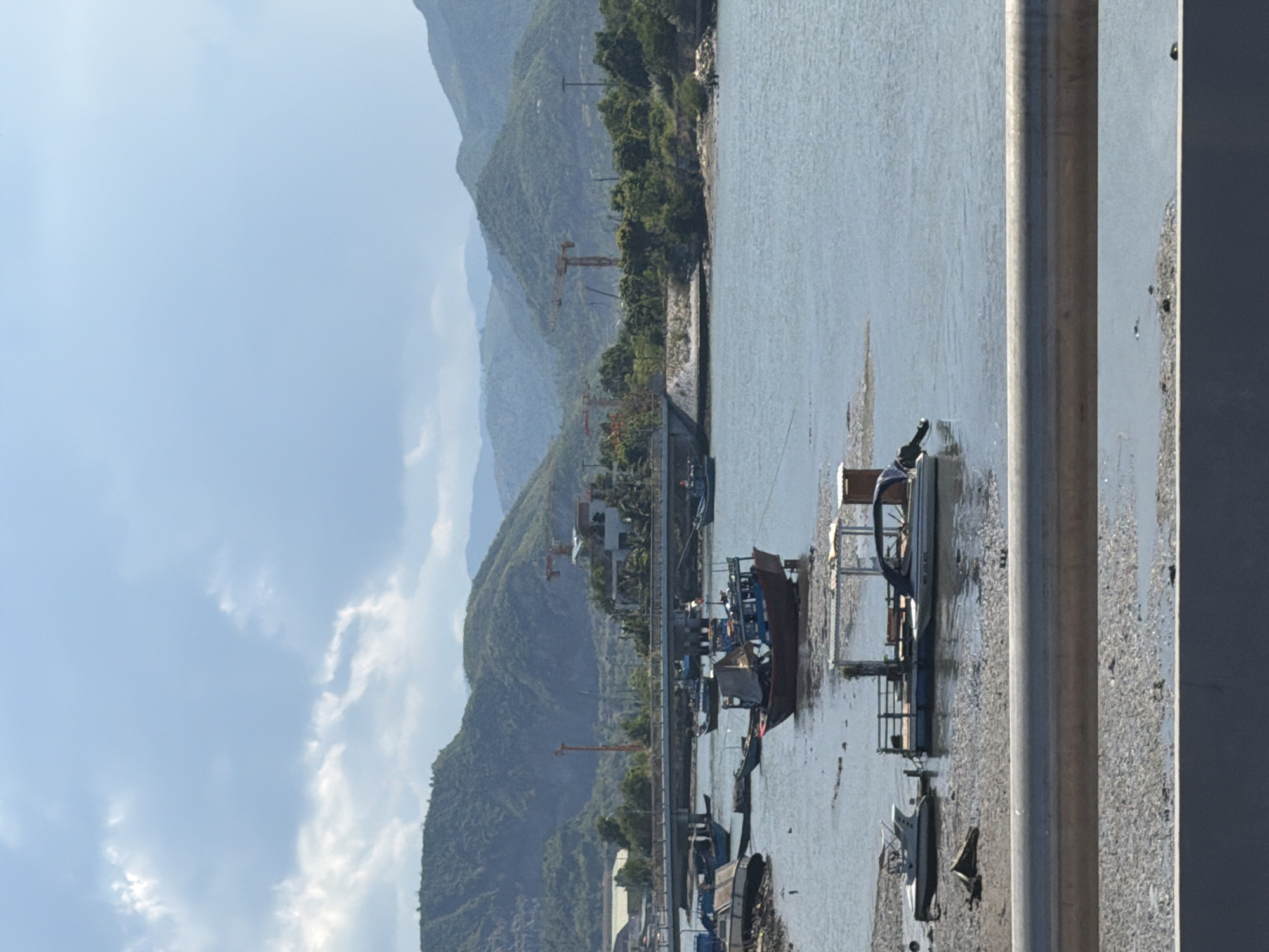 The image depicts a coastal scene in Nam Nha Trang Ward, Vietnam, on a bright, partly cloudy day. The foreground features a grey metal railing and a textured surface of mudflats, indicating low tide. In the middle ground, several boats are moored in the water or resting on the exposed mud. These include a large, dark red and blue fishing vessel with a weathered canvas cover, a smaller white motorboat with a blue canopy, and what appears to be a floating dock or pontoon structure with seating and a small shed. Further in the background, a bridge spans a body of water, with construction cranes and buildings visible on the distant shore. The landscape is dominated by lush green, rolling hills and mountains that recede into the haze. The sky is a vibrant blue with scattered white clouds. The overall impression is one of a working harbor or coastline, with elements of both traditional fishing and ongoing development. There are no visible people in the image.