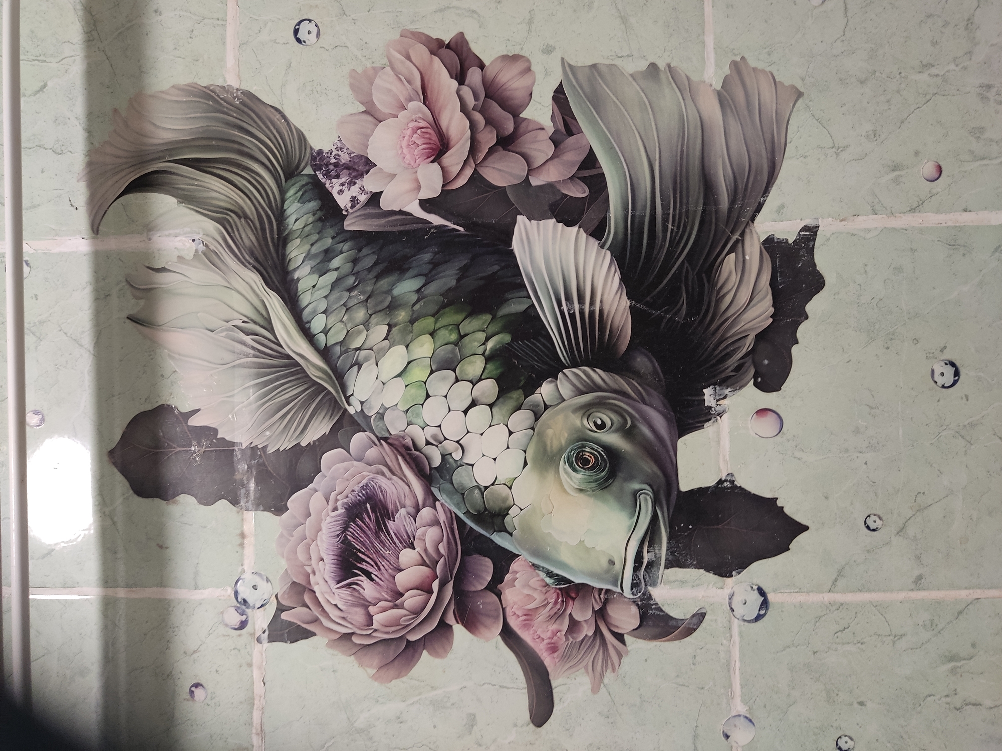 The image is a close-up of a decorative tile mural on a wall. The mural depicts a stylized koi fish surrounded by ornate flowers and leaves. The fish is predominantly green and silver with iridescent scales, and it has prominent fins and a large, expressive eye. The flowers are a soft pink and purple, with layered petals. Small, translucent bubbles are scattered across the tiles, adding a whimsical touch. The background is a light green, marble-patterned tile with visible grout lines. The lighting appears to be overhead, creating a slight glare on the surface of the tiles. This mural is located in the city of Noginsk, Russia.
