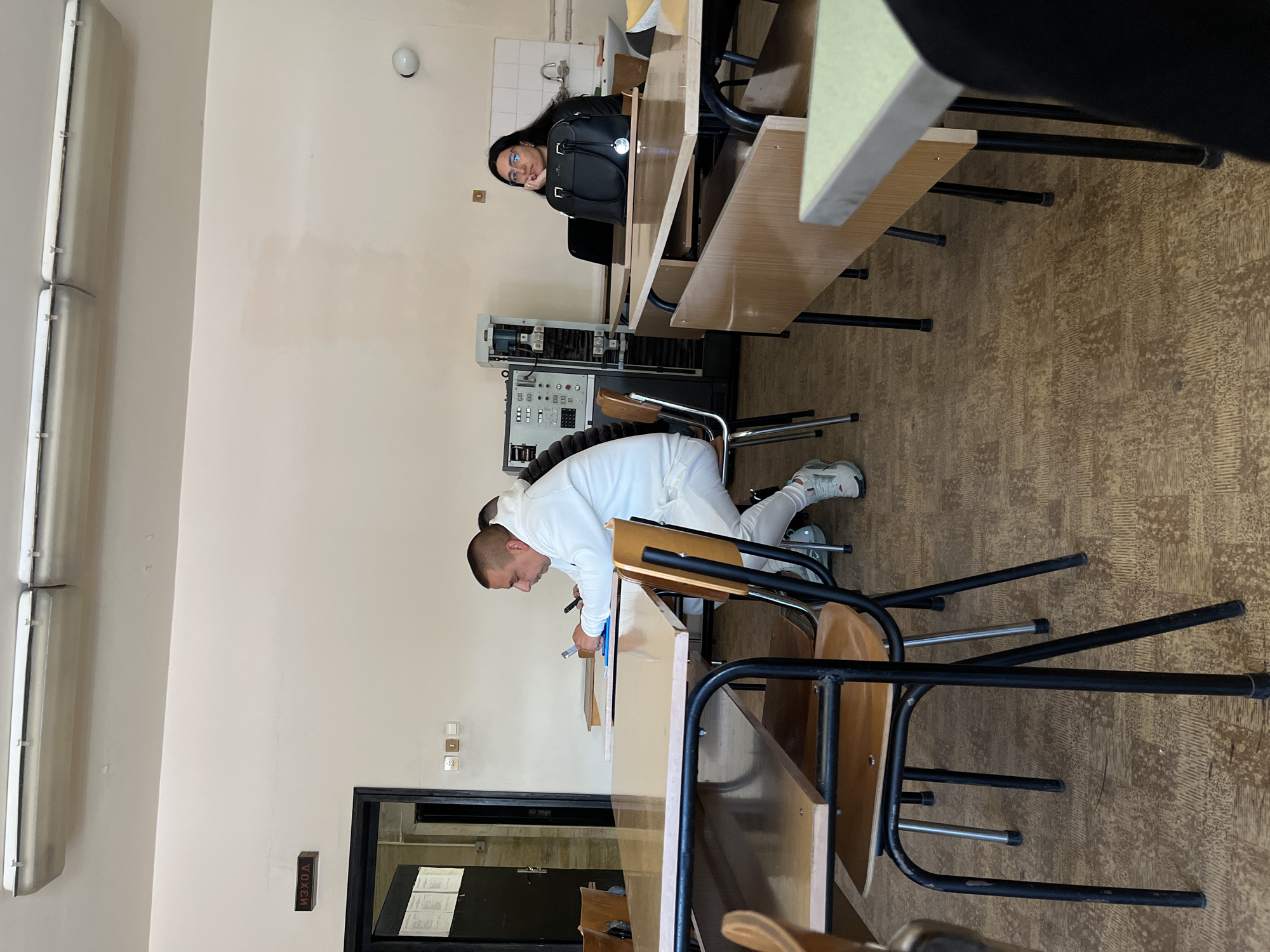 The image shows the interior of a classroom, likely a science lab, at the Plovdiv University "Paisii Hilendarski" in Plovdiv, Bulgaria. Two individuals are present. A woman with dark hair and glasses sits at a desk in the background, looking towards the camera. In the foreground, a man dressed in a white long-sleeved shirt and pants is leaning over a desk, appearing to write or draw in a notebook with a pen. Desks and chairs are arranged in rows. Lab equipment is visible against the wall. The walls are white with some markings. An exit sign is on the wall next to the door. A fluorescent light fixture is on the ceiling. The floor is covered with linoleum.