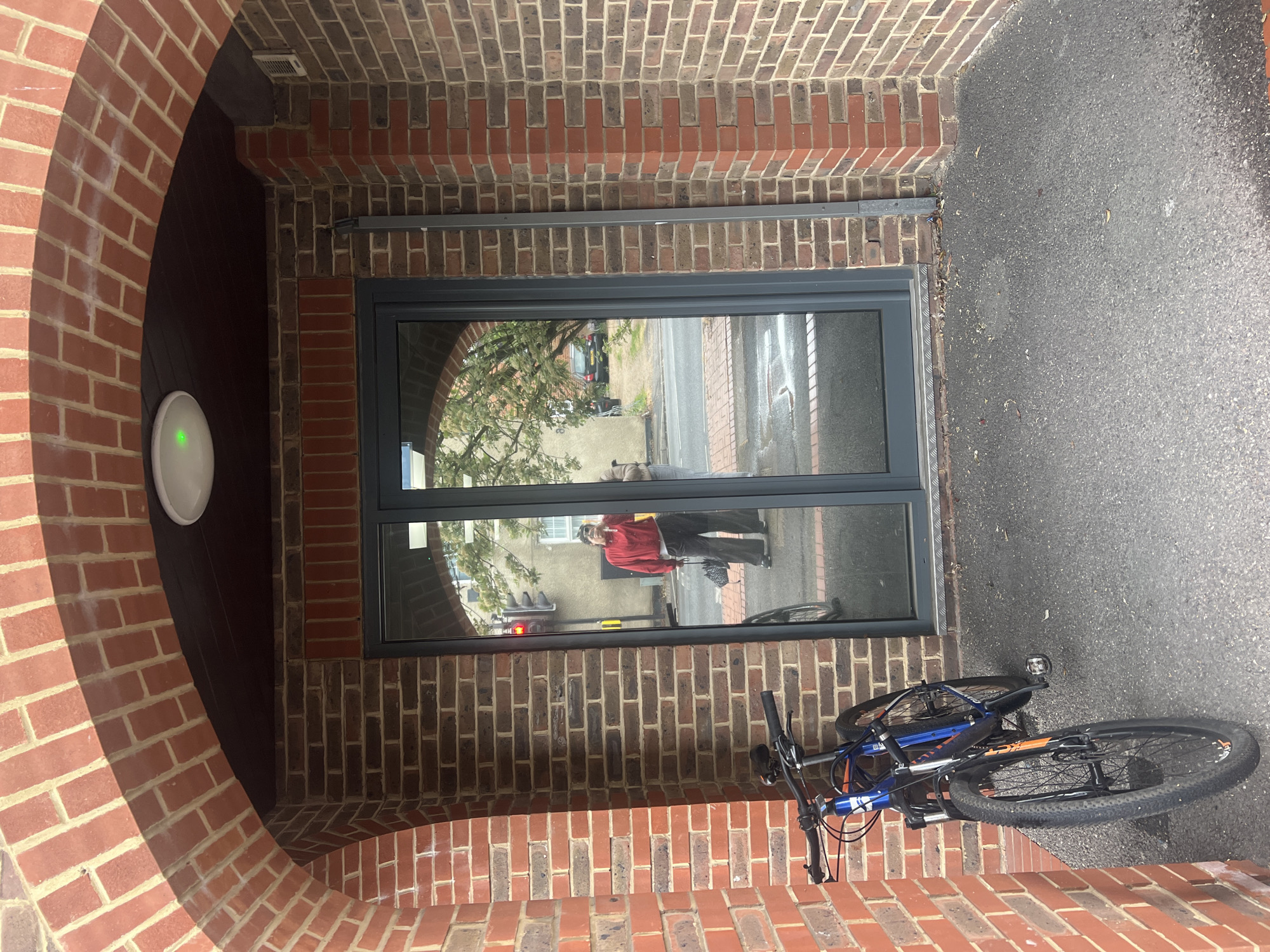 The image depicts a brick-lined entrance or archway in Mole Valley, United Kingdom. Within the archway is a modern gray-framed window reflecting a street scene. A person in a red jacket is visible in the window reflection. A blue and orange mountain bike lies on the gray asphalt outside the entrance, partially obscuring the view of the street. On the left wall of the archway, there is a white circular device with a green light. The brickwork is primarily red, with some areas featuring darker, almost brown bricks.