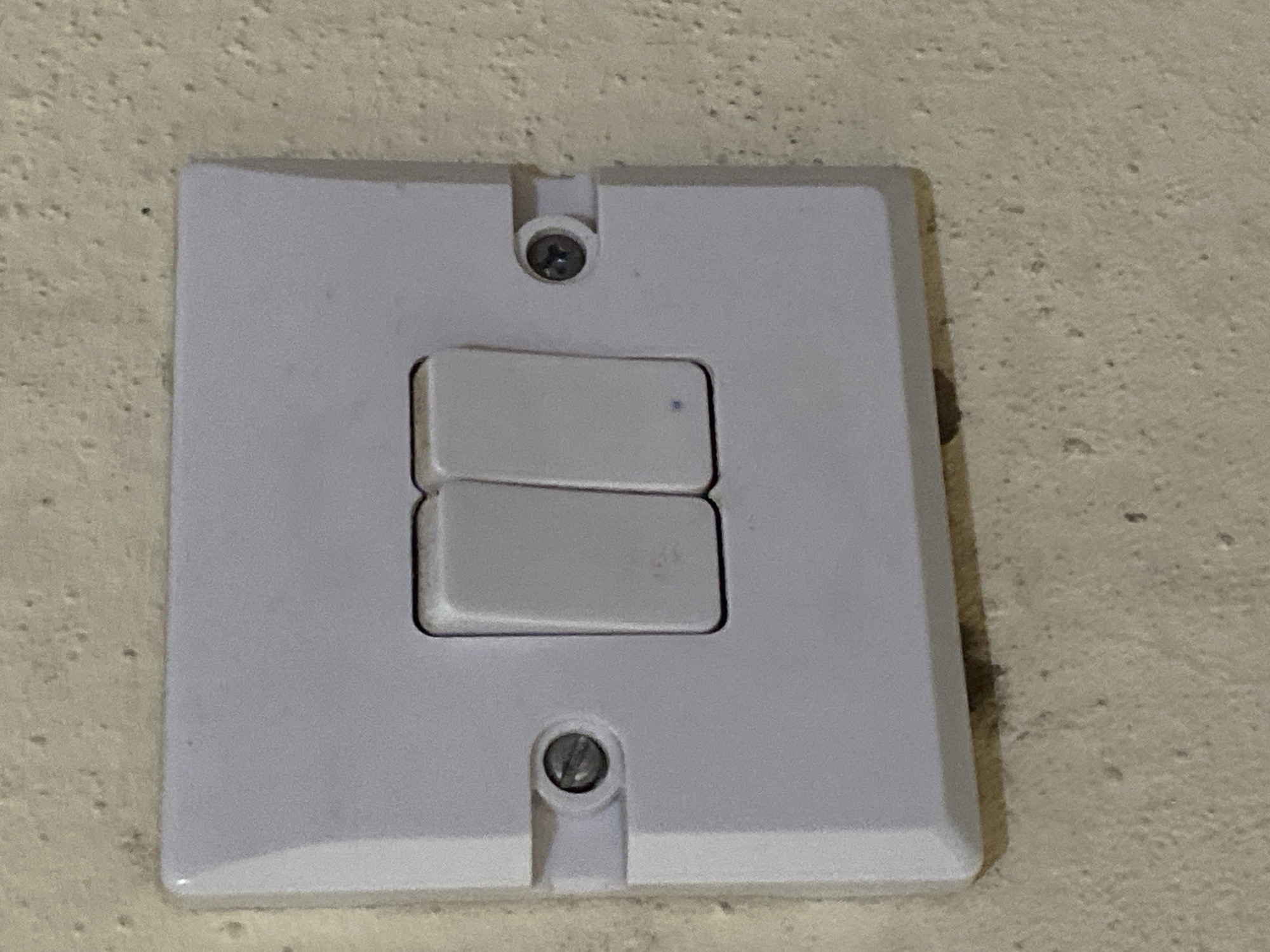 This media file contains a close-up, eye-level shot of a white double light switch mounted on a textured, light-colored wall. The switch plate is square with rounded corners, and the two rocker-style switches are stacked vertically. The upper switch is in the "off" position, while the lower switch appears to be in the "on" position, indicated by a small blue dot on its surface. Two screws are visible, one above the upper switch and one below the lower switch, securing the plate to the wall. The wall texture is rough and uneven, with small dark specks embedded in the material. There is no visible text in the image. The lighting suggests an indoor setting, possibly during the day given the even illumination. The scene appears to be a common domestic or commercial setting in Jauro Yinu, Nigeria. No people or specific activities are depicted, only the static object and its environment.