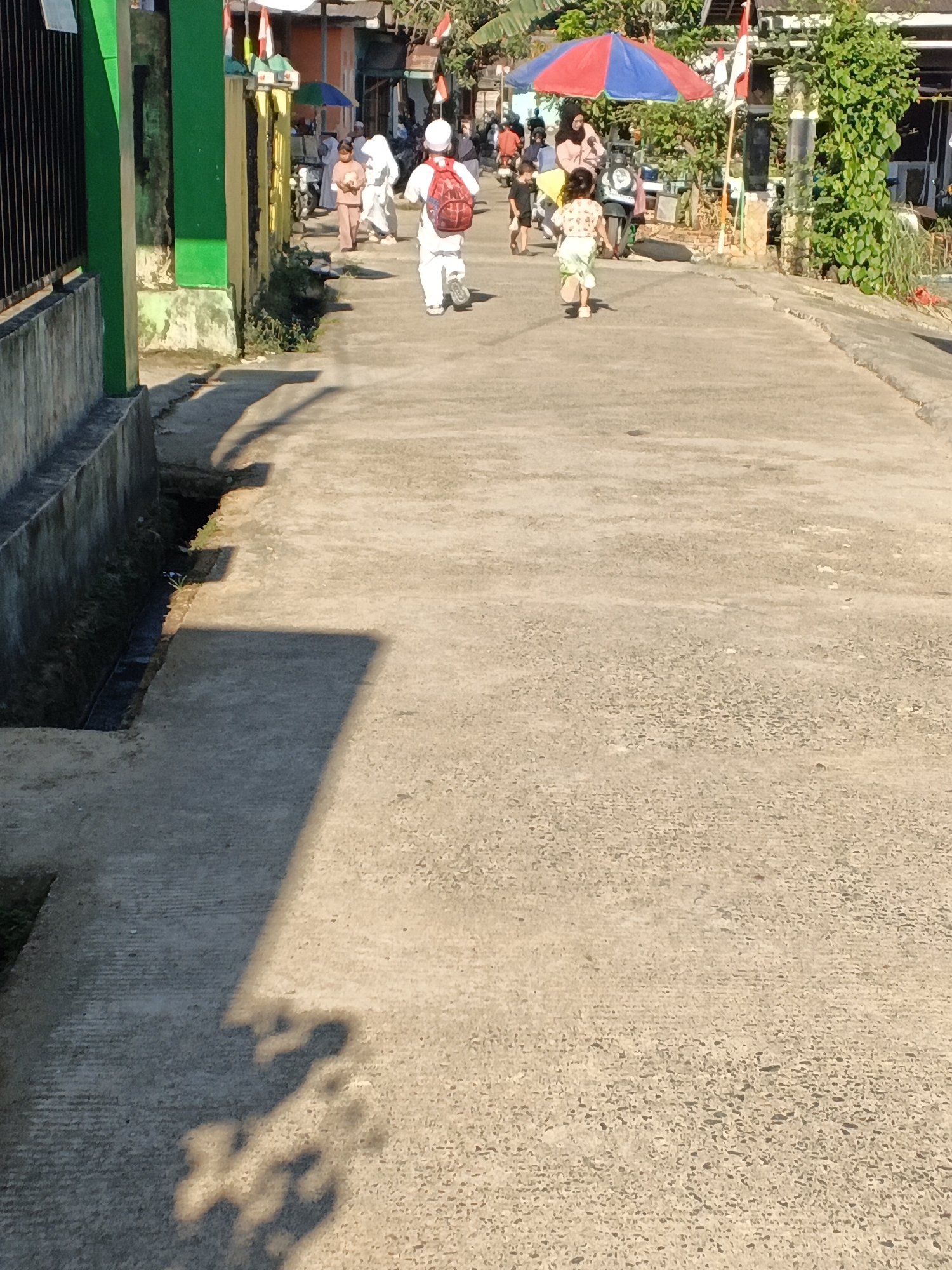 The image shows a street scene in a village. In the foreground, a child runs away from the camera. The child is wearing a light-colored shirt and pants. Further down the street, a person in a white robe and backpack walks towards the camera. Other people are seen in the background, some of whom appear to be children. There is a red and blue umbrella on the right side of the street. The street is paved and lined with buildings and greenery. The scene appears to be in a rural or suburban area.