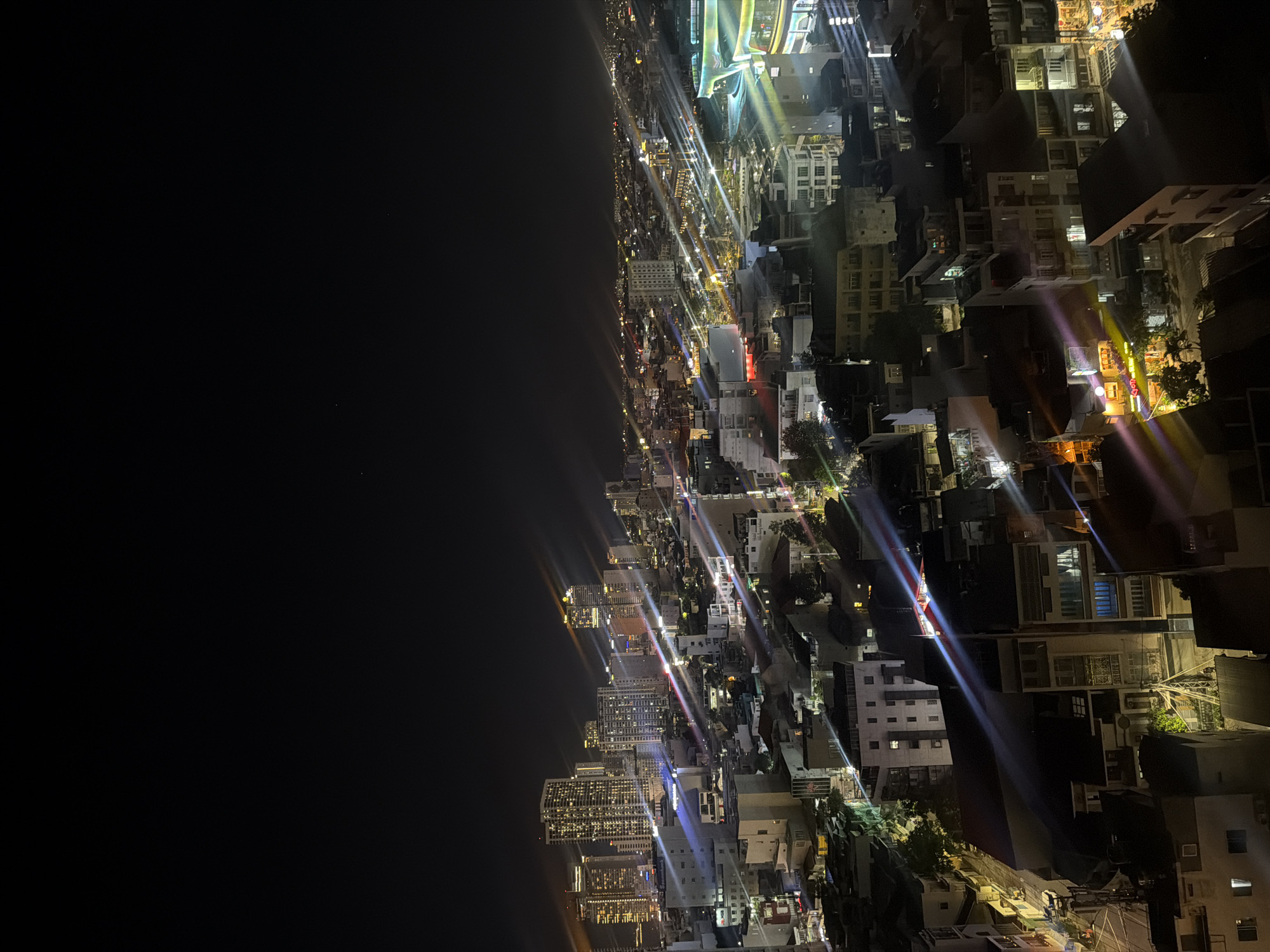 An aerial nighttime shot captures the urban landscape of Nha Trang Ward, Vietnam. The scene is dominated by a dense cluster of buildings, ranging from low-rise structures to towering skyscrapers, all illuminated by artificial lights. Streaks of light, likely from streetlights and building illuminations, are dispersed across the image, creating a vibrant, almost abstract effect against the dark night sky. The architecture appears to be a mix of modern and older styles, with various building heights and designs visible. The city stretches into the distance, with a high concentration of lights suggesting a bustling urban center. The sky above is a deep, inky black, with a few faint stars visible in the upper portion of the frame. The overall impression is one of a lively and dynamic city at night, seen from a high vantage point.