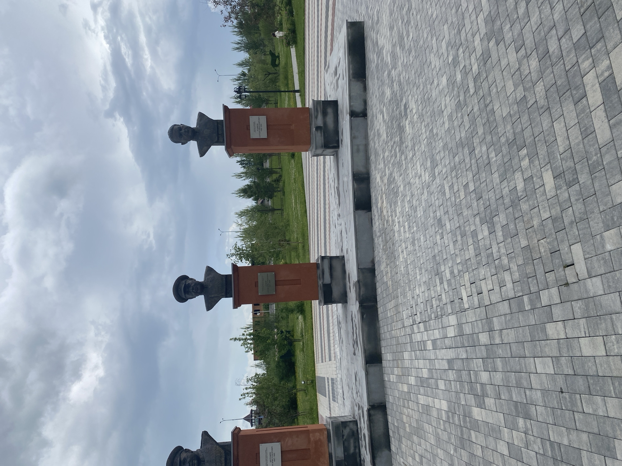 The image presents an outdoor memorial space, identified as the Alley of Heroes within the Square of Memory (Сквер Памяти) in Tyumen, Russia, under an overcast sky. The foreground features a wide paved area composed of rectangular interlocking pavers in shades of grey. The paving patterns vary, with some sections displaying a mixed-grey block arrangement and others featuring parallel stripes of alternating light and dark grey blocks.
Three bronze busts of male figures are prominently displayed on individual reddish-brown, rectangular pedestals, which in turn are mounted on multi-tiered, dark grey bases. From left to right:
1. The leftmost bust is partially visible, depicting a man in a military uniform with a peaked cap. Its plaque is obscured.
2. The central bust is a depiction of Onufriy Khristoforovich Kashtanov (1893-1943), shown in military uniform with a peaked cap. A white rectangular plaque affixed to the pedestal identifies him by name and life dates in Cyrillic.
3. The rightmost bust is a depiction of Sergey Vladimirovich Kuzhelny (1960-1995), portrayed without headwear, possibly in a service uniform or civilian attire. A white rectangular plaque on this pedestal displays his name and life dates in Cyrillic.
Behind the busts, a landscaped park area extends into the mid-ground and background. This area comprises green lawns, various deciduous and coniferous trees, and a paved pathway winding through the greenery. On the right side of the park, a green animal-shaped topiary sculpture (resembling a deer or horse) stands near the pathway, where a small child wearing light-colored clothing is walking away from the viewer. Several streetlights are visible among the trees. In the far background on the left, an unfinished building structure is partially visible above the tree line. The sky is predominantly covered with grey and white stratocumulus clouds, indicating an overcast day.