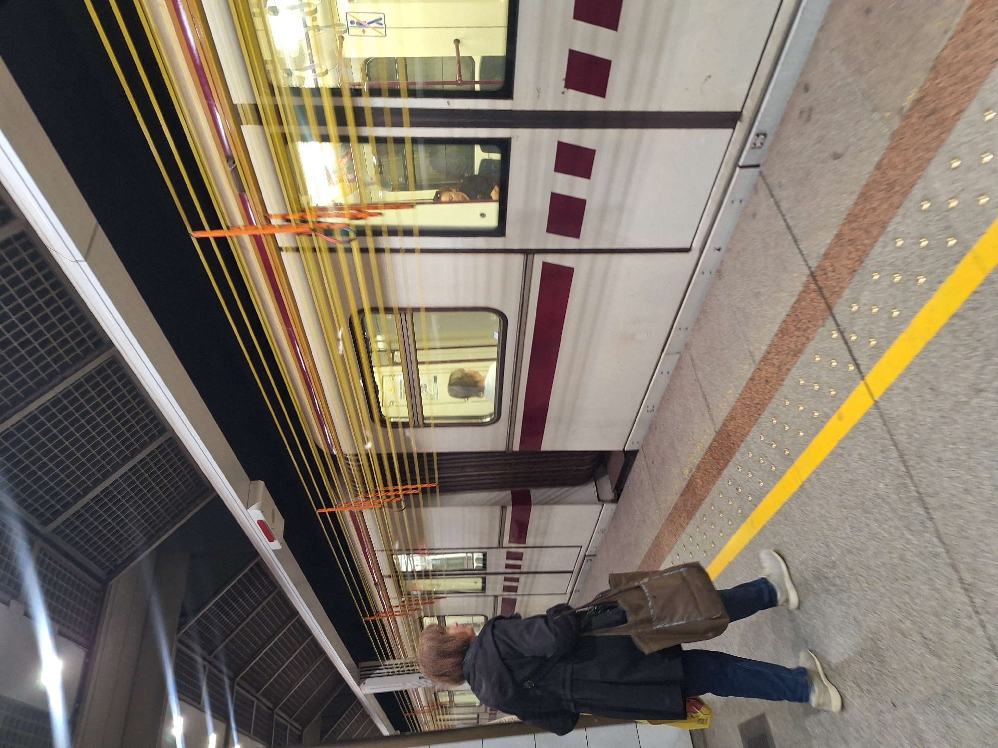 A person is standing on a train platform next to a train. The train has a white exterior with burgundy stripes and a few small maroon square patterns. The person wears a dark coat, jeans, and white shoes, and has a tan tote bag. A yellow line marks the edge of the platform. The train station has a modern architecture with a metal grid ceiling and yellow support cables. The location is Sofia, Bulgaria.