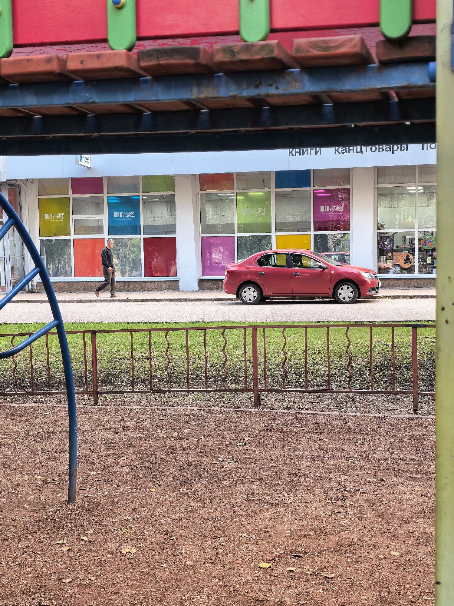 A man walks along the sidewalk in front of a store with colorful windows. A red car is parked on the street. The scene is in a city in Russia, likely Sterlitamak, as suggested by the Russian text on the store awning, which reads "КНИГИ канцтовары ПО" ("Books, stationery, etc."). The foreground shows a playground with a red gravel surface and a blue metal structure. A brown metal fence separates the playground from the sidewalk. The overall impression is of a public space with a mix of commercial and recreational elements.