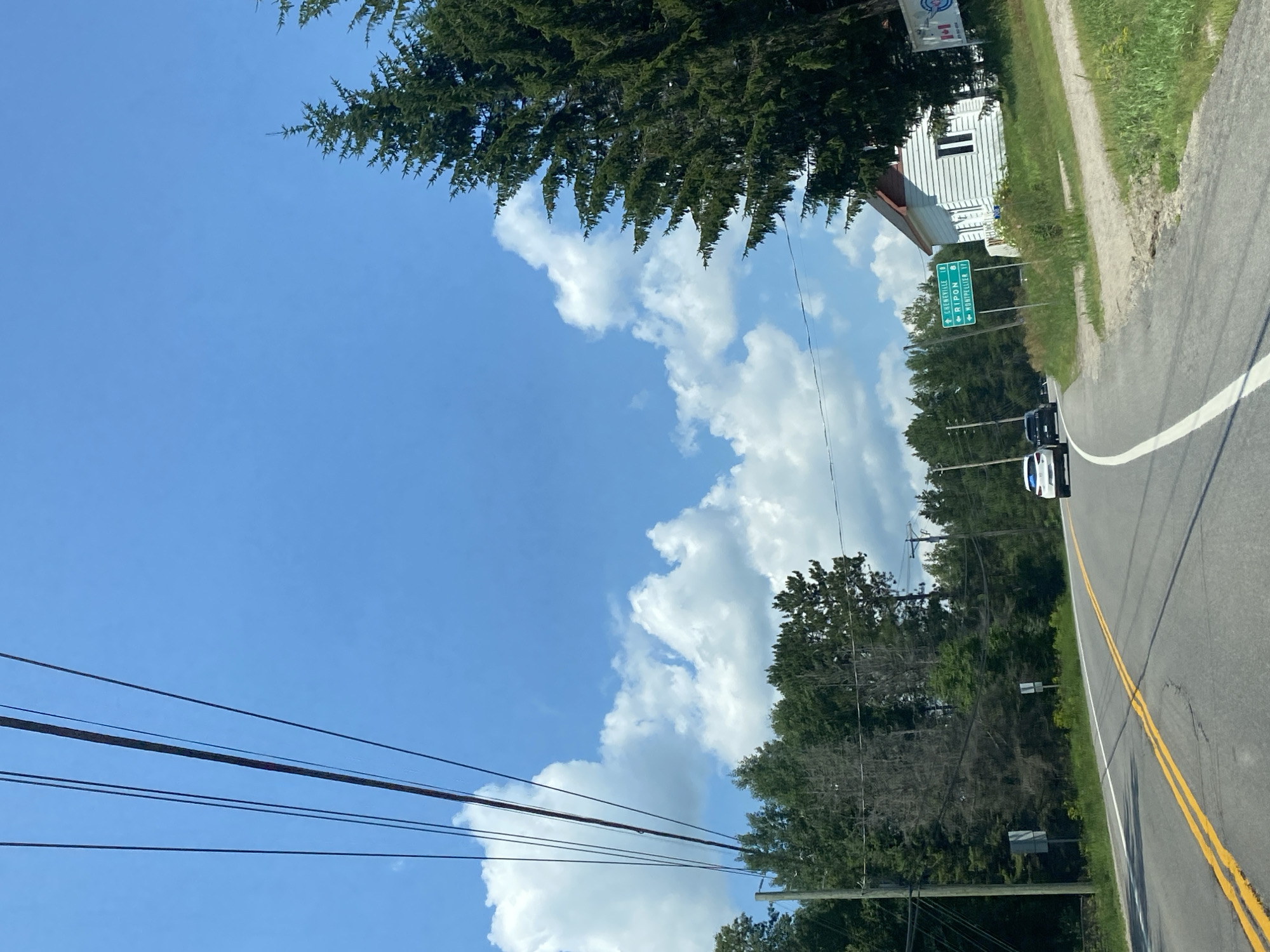 The image depicts a rural road in Saint-André-Avellin, Canada, on a sunny day with a blue sky and scattered clouds. A paved road with yellow double lines curves to the right. On the left side of the road, there are power lines strung across the frame and a dense line of evergreen trees. On the right side of the road, there is a small white house with a red roof, surrounded by grass and trees. A green road sign indicates directions to "CHENNEVILLE 10", "RIPON 8", and "MONTMELIANT 17". Two cars, a white SUV and a black sedan, are driving on the road, heading in the same direction as the viewer. The overall scene suggests a quiet, scenic drive through a wooded area.