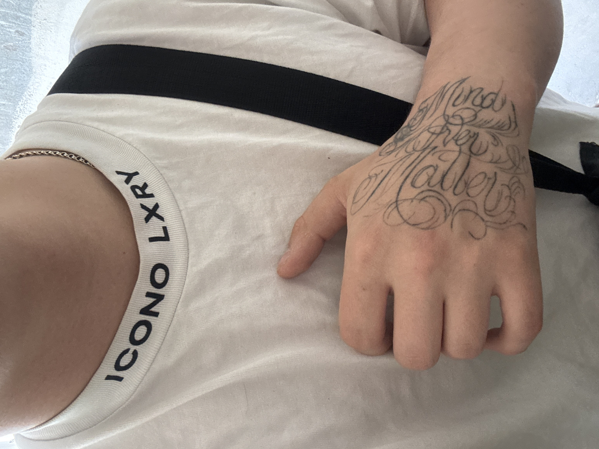 A close-up photograph depicts a portion of a person's torso and hand, lying in an unknown location. The person is wearing a white t-shirt with a black band across the chest and the words "ICONO LXRY" vertically printed on the collar. The person's left hand rests on the t-shirt, displaying a tattoo on the wrist and back of the hand with the words "Mind Over Matter" in elaborate script. A thin gold chain is visible around the neck. The background appears to be a light-colored surface.