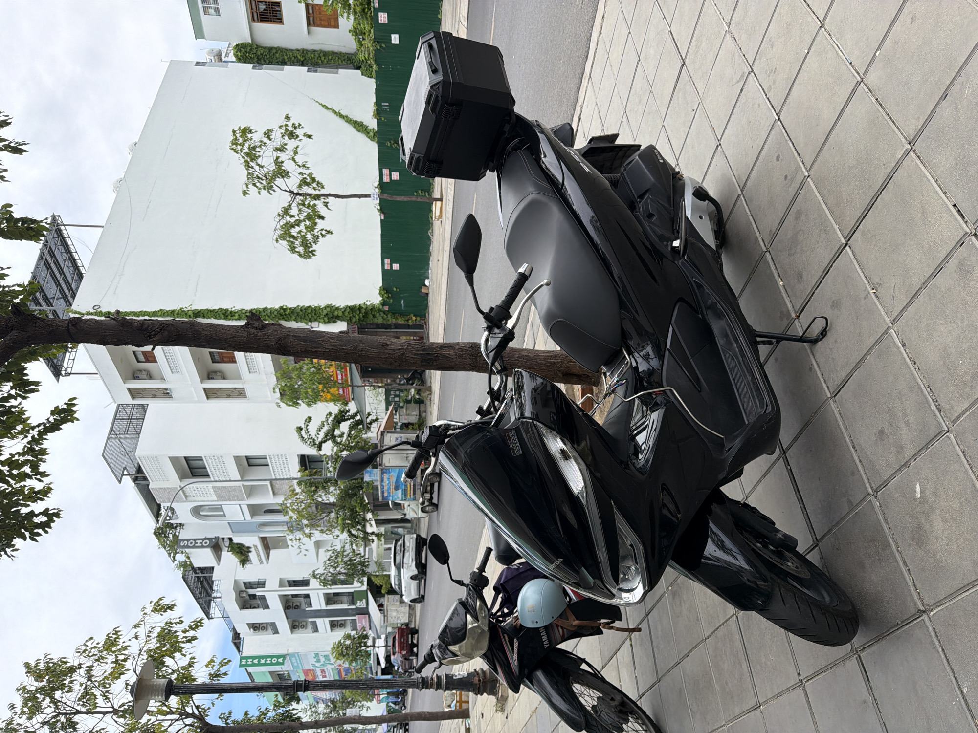 The image depicts a street scene in Nha Trang, Vietnam. In the foreground, two black motorcycles are parked on a tiled sidewalk. The motorcycle on the right is a scooter with a large black storage box mounted on the rear. A light blue helmet is attached to the handlebars of the motorcycle on the left. Behind the motorcycles, a street runs parallel to the sidewalk, with several cars and a truck visible. In the background, white buildings with multiple stories line the street. Green trees and foliage are present throughout the scene, with some vines climbing the sides of the buildings. The sky is overcast.