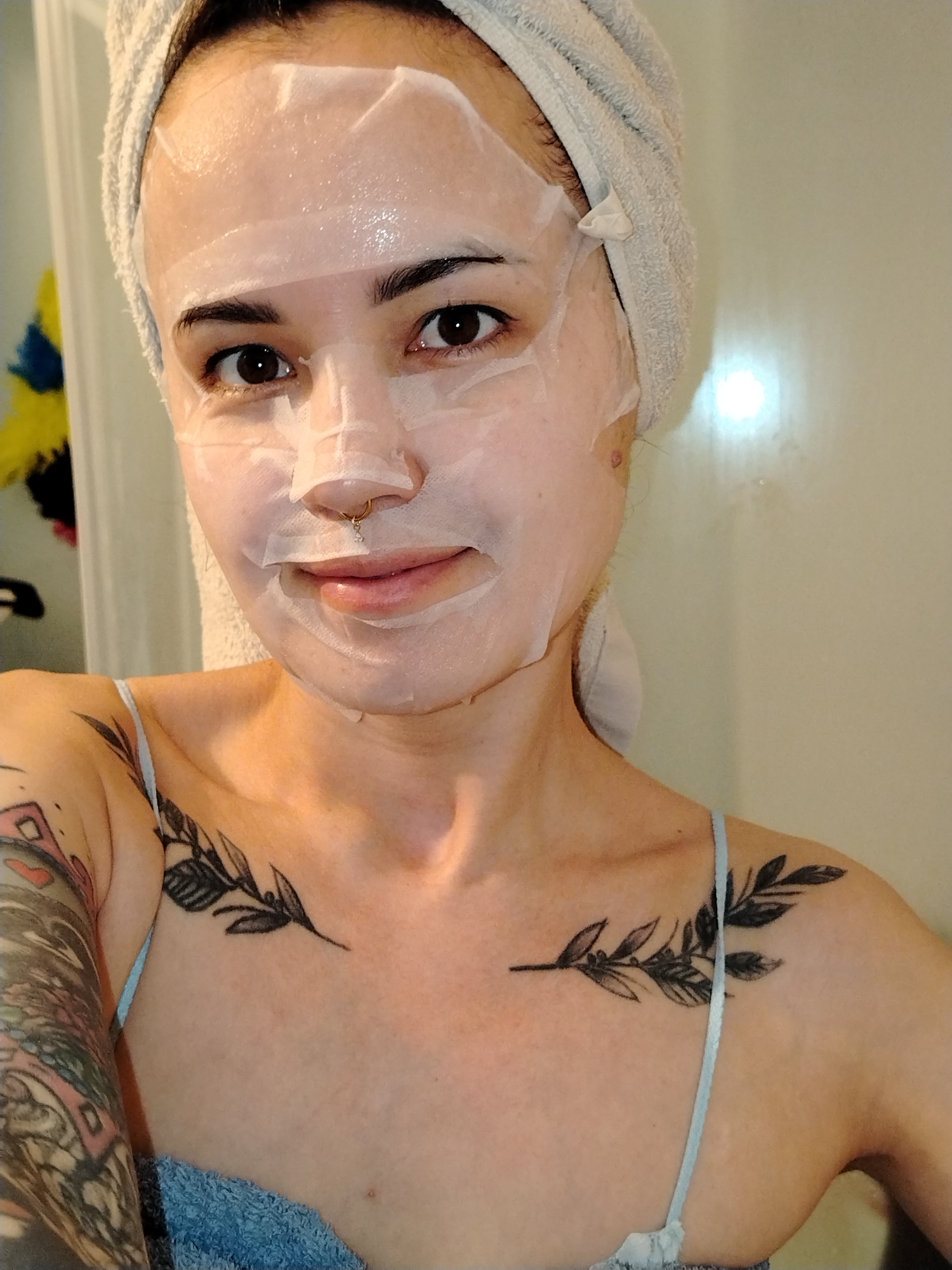 A woman is facing the camera, wearing a sheet mask on her face and a towel wrapped around her head. She has a nose ring and visible tattoos on her left arm and chest. The background is a light-colored wall.