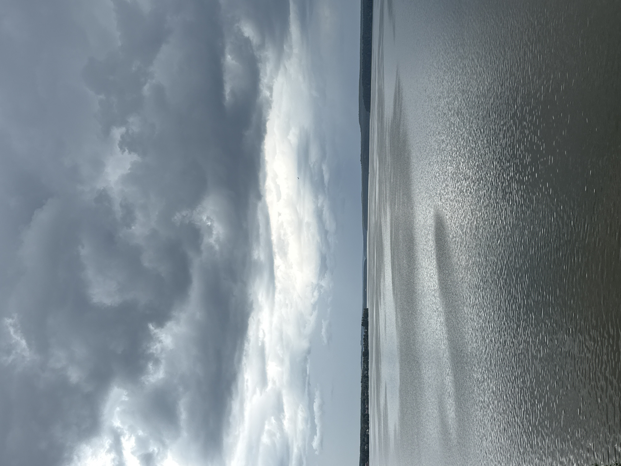 An overcast sky hangs heavy over a wide, calm body of water. Dark, heavy clouds dominate the upper portion of the image, with lighter patches suggesting a break in the storm. The water reflects the muted light, creating a textured surface of ripples and gentle waves. Along the distant horizon, a dark, tree-lined shore is visible, hinting at a natural landscape surrounding the water. The scene appears to be a natural body of water, possibly a lake or a wide river, in Votkinsk, Russia. There are no people or specific objects readily identifiable within the frame. The overall atmosphere is somber and tranquil.
