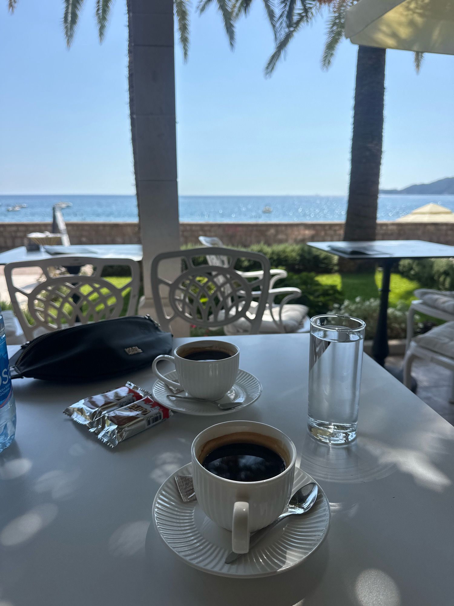 This is a close-up, eye-level shot of a white table on a sunny day with a view of the sea. Two white cups of coffee with saucers and spoons are placed on the table, along with two individually wrapped snack bars, a black clutch purse with "KARL" written on it, and a glass of water. A partially visible bottle of water is to the left. In the background, white outdoor chairs and tables are visible, along with palm trees and a bright blue sky. The sea, with some boats, stretches to the horizon. The scene is likely set in Rafailovići, Montenegro, a coastal town known for its beaches and outdoor dining.