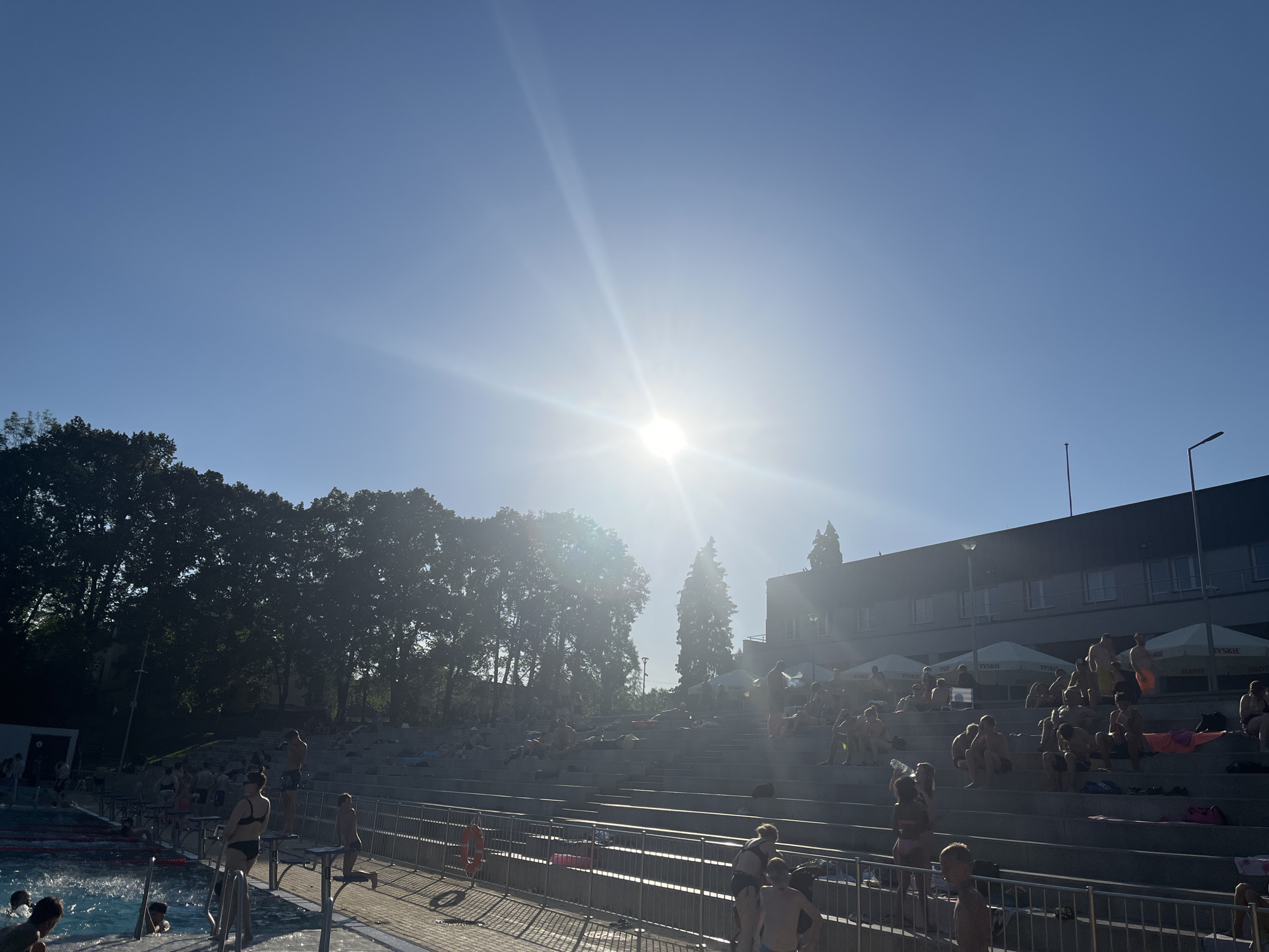 This is a wide shot of an outdoor swimming pool complex on a sunny day. The sun is high in the sky, creating a bright glare and lens flare. The scene is filled with people, most of whom are in swimsuits, enjoying the warm weather.
The primary subjects are the numerous people scattered throughout the complex. Many are relaxing on tiered, concrete grandstands that overlook the pool area. Some are lying down on towels, while others are sitting and socializing. There are also people in and around the pool itself, either swimming or standing on the edge. Several individuals are visible on diving boards.
The setting appears to be a public swimming facility, with a large pool visible on the left side of the frame. The tiered seating is a prominent feature, designed to accommodate many spectators or sunbathers. In the background, there is a building with multiple windows and a flat roof, possibly a changing facility or administrative area. Trees line the perimeter of the complex, suggesting a natural or park-like environment surrounding the man-made structures.
The activity taking place is clearly leisure and recreation. People are swimming, sunbathing, and spending time outdoors. The atmosphere seems relaxed and summery, with the bright sunlight contributing to a feeling of warmth and enjoyment.
Notable details include the clear blue sky, indicating fair weather. The strong sunlight suggests it is likely midday or early afternoon. There is a safety railing with life preservers visible near the pool. Some umbrellas are set up on the grandstands, providing shade. Visible text includes "TYSKIE" printed on white umbrellas.