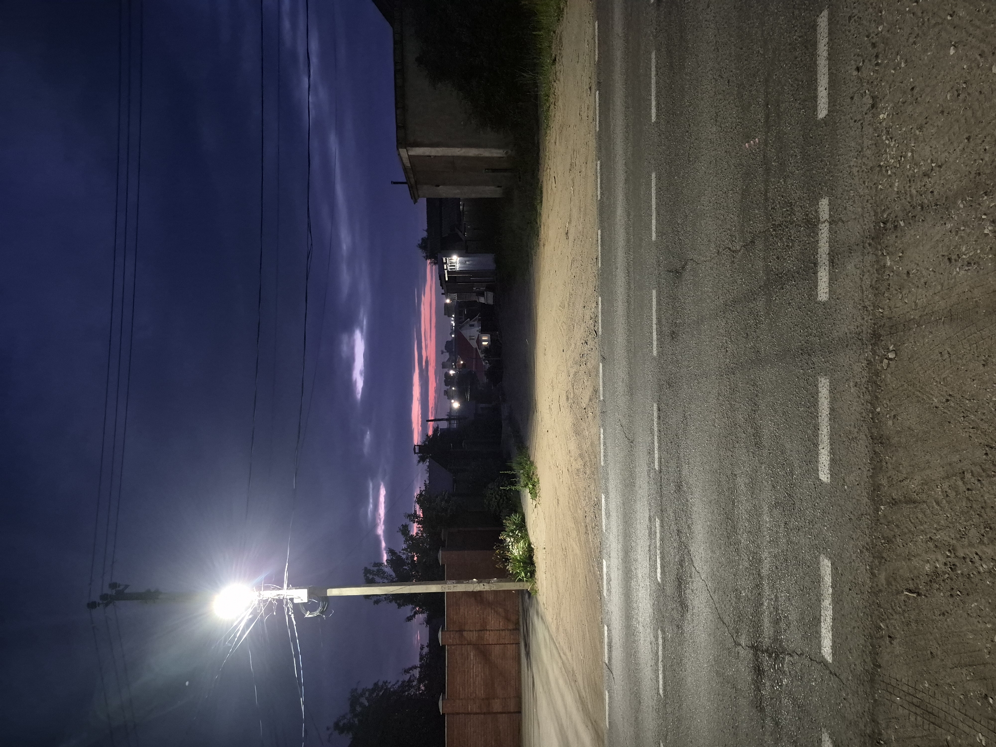 The image depicts a street scene at dusk in Chaikovsky, Russia. A street lamp illuminates a paved road with white dashed lines. To the left, a red brick fence lines the edge of the property. Beyond the fence, trees and the sky are visible. In the center of the image, a dirt path leads towards a residential area with houses and buildings. The sky is dark blue with purple and pink hues near the horizon, indicating the end of daylight. The overall atmosphere is quiet and still.