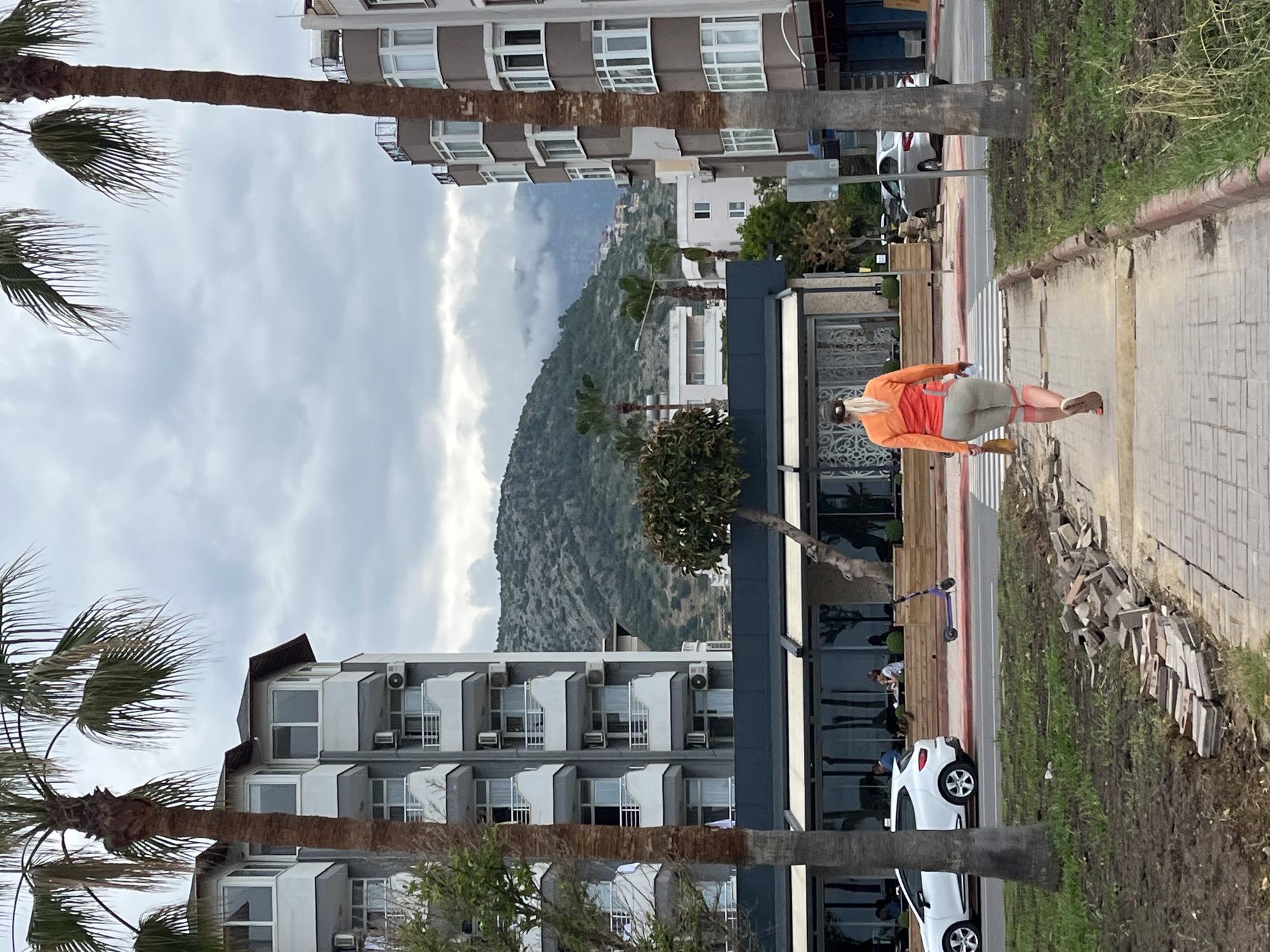 A woman in an orange jacket and light-colored pants walks across a crosswalk in Alanya, Turkey. She holds a brown bag in her left hand. Palm trees frame the view. A mountain rises in the background, partially obscured by clouds. Buildings, including multi-story hotels or apartments, line the street. A restaurant with large windows and a dark facade is visible. Cars and a scooter are parked along the street. Discarded or unused paving stones are piled up next to the sidewalk.