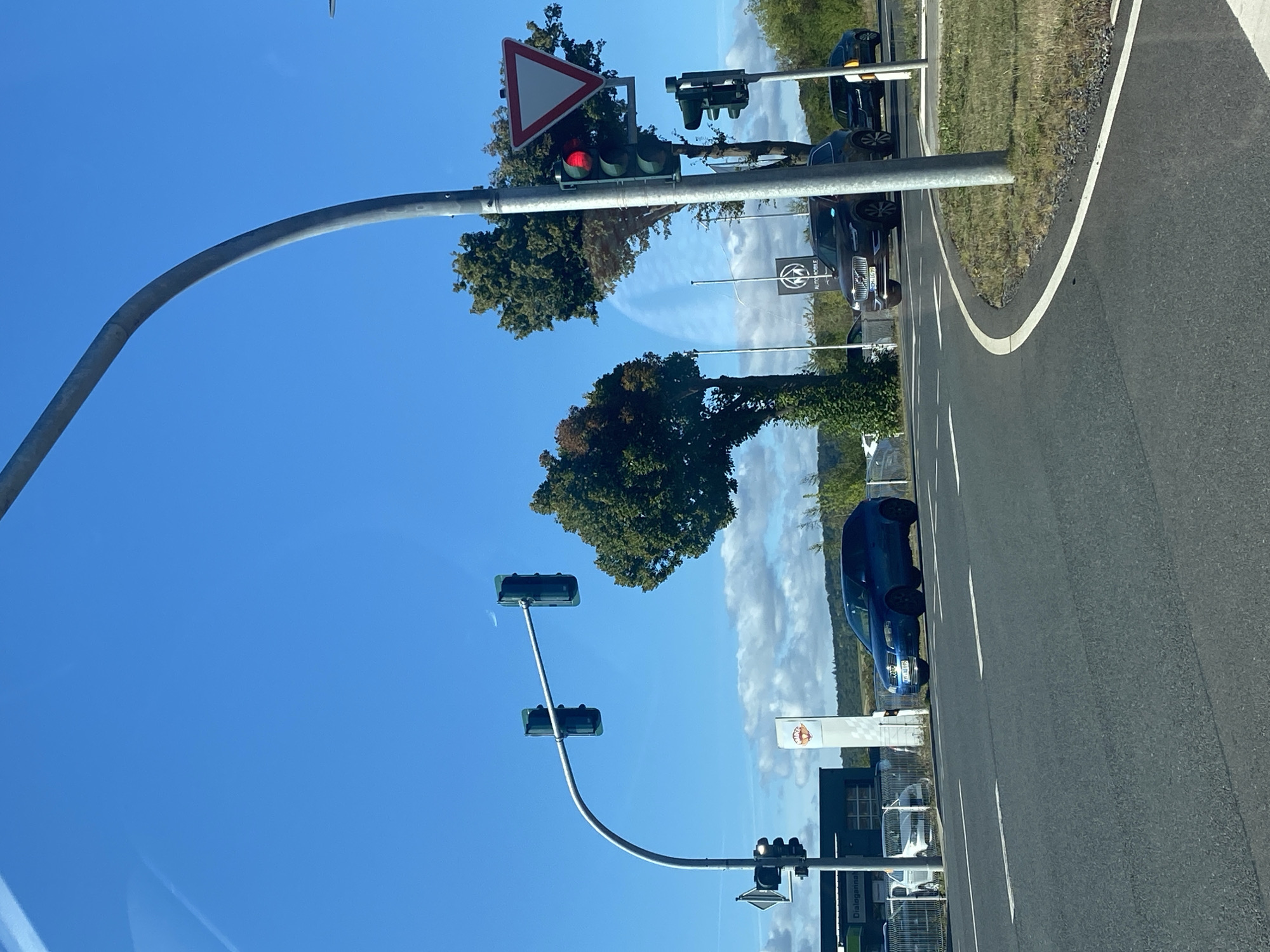 The image captures a road intersection in Werther, Germany, under a clear blue sky with scattered clouds. A traffic light on a curved pole shows a red light illuminated. Nearby, a give way sign is visible. On the road, a dark-colored SUV is stopped, with another car partially visible behind it. On the left side of the road, a blue hatchback is also stationary. In the background, buildings with signage indicating businesses such as "Dialogkanal" and "Auto MOBILE" are present, along with several parked vehicles. Trees are scattered throughout the scene, contributing to the outdoor environment. The road itself features lane markings, and a grassy area borders the road. The perspective suggests the image was taken from a moving vehicle.