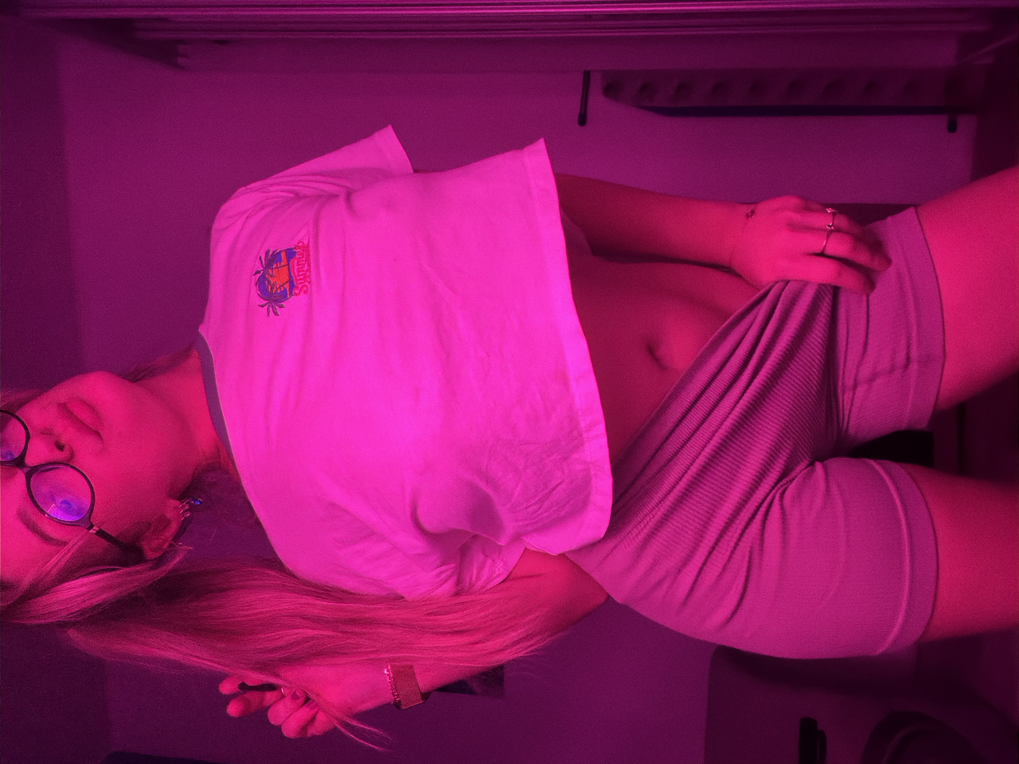 A young woman with blonde hair and wearing glasses is lying down. She is wearing a light-colored t-shirt with a "Summer" logo on it and matching shorts. The lighting is predominantly pink. The location is assumed to be in an indoor space in Большая Шильна, Russia, based on the provided context. The woman is holding her hand on her stomach area. The background shows parts of what appear to be light fixtures or architectural details.