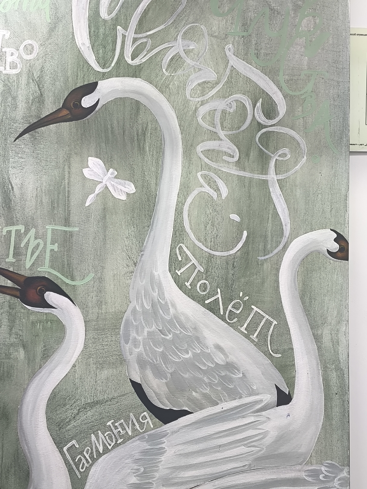The image is a close-up of a painted artwork featuring three swans against a mottled green and grey background. The swans are painted in shades of white and grey, with brown and black details on their heads and beaks. One swan is prominently centered, with two others partially visible on either side. A white dragonfly is depicted floating near the central swan.
Text is written in various places around the swans. Some text is in a looping, cursive script in white, while other text is in a sans-serif font in a pale green color. The word "Гармония" (Harmony) is written near the bottom left in white, and "Полеём" (Let's Fly) is partially visible in white on the right. The location of the image is not determinable from the provided image.