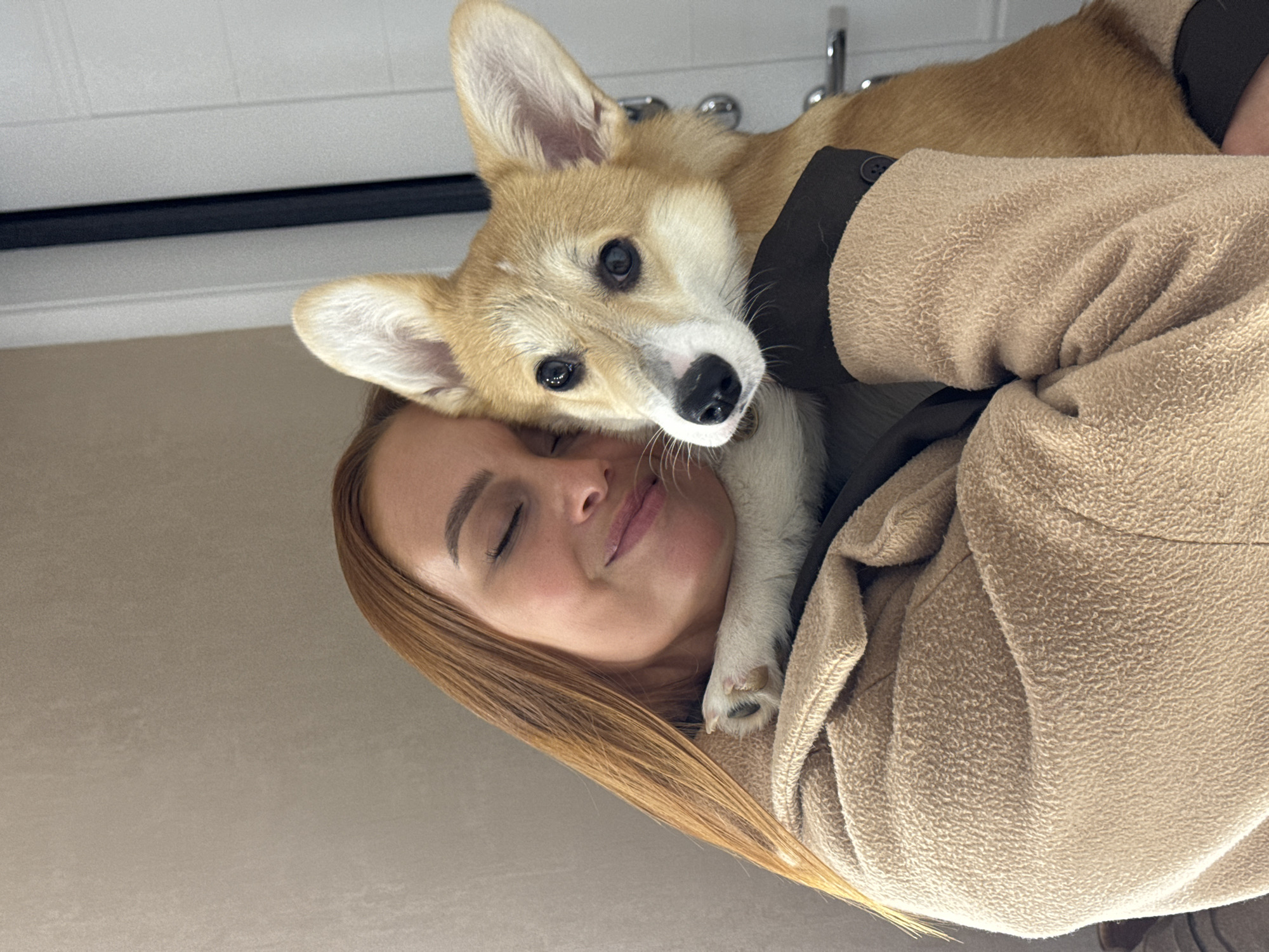 The image captures a close-up, vertically oriented portrait of a woman and a corgi dog in an indoor setting in Chaikovsky, Russia. The background is softly blurred, showing light-colored walls or cabinets and hints of metallic fixtures, possibly from a sink area.
On the left, a woman with long, straight, reddish-brown hair has her eyes closed, displaying a peaceful and gentle smile. She is wearing a textured, light beige or tan jacket, which appears soft and warm. Her head is tilted slightly, resting as if in comfort.
Nestled against her, with its head resting on her shoulder and cheek, is a small corgi dog. The dog has typical corgi markings with light brown fur and white on its face, chest, and paws. Its dark, expressive eyes are wide open, looking directly at the viewer. A small black nose and white muzzle are prominent. One of the dog's front paws, with visible claws, rests gently on the woman's shoulder. The dog also appears to be wearing a dark harness or undershirt beneath the woman's jacket.
The photograph conveys a strong sense of affection, warmth, and close companionship between the woman and her pet. The lighting is soft and natural, suggesting it is daytime. There is no visible text in the image.