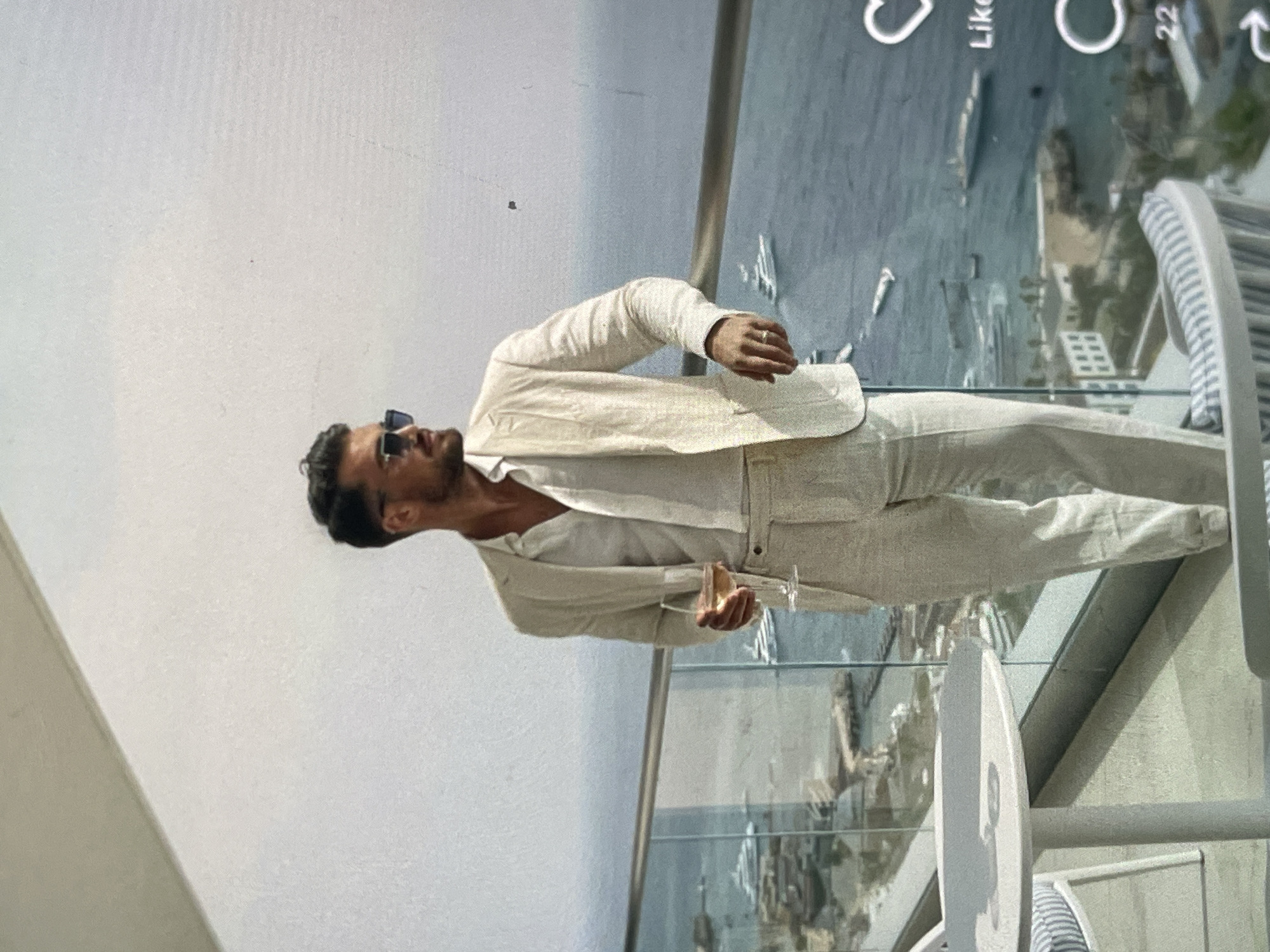 A man dressed in a light-colored linen suit and white shirt stands on a balcony, holding a glass of rosé wine. He wears sunglasses and a ring on his left hand. The balcony overlooks a city and a body of water with several boats. A small round table and chairs are present on the balcony. The sky is overcast.