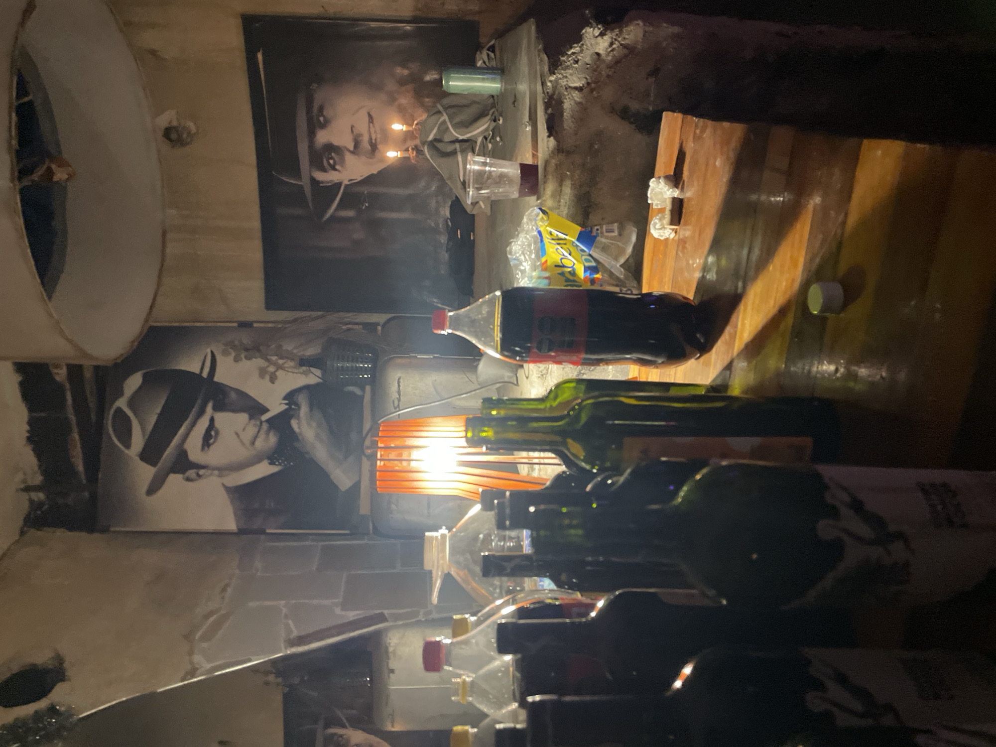 The interior scene depicts a dimly lit space, likely a basement or cellar, cluttered with objects. Two black and white portraits of men in hats are visible on the wall. A Coca-Cola bottle and several wine bottles are prominent in the foreground, illuminated by an orange lamp. A wooden surface is present, with items scattered on it, including what appears to be a plastic cup with a dark liquid and crumpled white material. The environment appears unkempt and dimly lit, suggesting a casual, perhaps neglected space. There is no visible information confirming the location in Buenos Aires, Argentina.