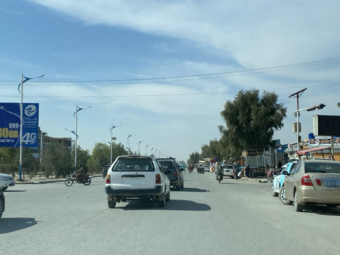 This image captures a wide-angle street view in the city of Kandahar, Afghanistan, under a clear to partly cloudy daytime sky. The perspective is from within a moving vehicle, looking forward down a multi-lane asphalt road.
In the immediate foreground, the back of a white station wagon with an exposed roof rack is prominently centered, facing away from the viewer. To its right, a gold-colored sedan with a blue license plate is parked alongside the road, partially obscured by a light blue sedan parked slightly in front of it. On the far left, a portion of a white vehicle's front is visible.
Traffic is moderately heavy, with several cars and at least two motorcycles visible further down the road. One motorcyclist, wearing dark clothing, is visible mid-left, heading away from the viewer.
The road is lined with distinctive streetlights featuring blue, wing-like accents. Overhead power lines crisscross the sky. On the left, a large blue billboard for "Salaam" telecommunications advertises "999 80GB 4G" mobile data. Beyond the billboard and road median, some residential or commercial buildings are visible.
To the right, a row of commercial establishments with various signs and awnings borders the road. Pedestrians are visible near these shops. A tall pole on the right side of the road features what appears to be a solar panel and a dark rectangular sign or display. Mature trees with dense green foliage are scattered along both sides of the road, with a notable cluster on the right midground. The overall scene depicts a busy urban thoroughfare.