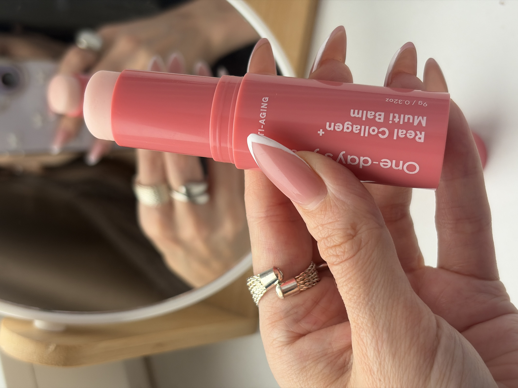 A close-up photograph displays a left hand holding a coral-pink "One-day's you Real Collagen + Multi Balm" stick, which is partially extended to reveal a lighter pink product. The hand exhibits manicured nails with a natural pink base and white tips. A silver-colored, open-ended cuff ring with a textured surface is worn on the ring finger. In the background, a circular mirror with a light wooden frame reflects a partial image of the same individual, holding a smartphone with a light-colored, patterned case, indicating a self-recording or photography action. The reflected hand also wears a silver-colored ring on the ring finger. The immediate environment is an indoor setting, likely a vanity or bathroom, with a white surface visible at the base. The image contains no visual cues or landmarks to identify a specific location within Saint Petersburg, Russia.