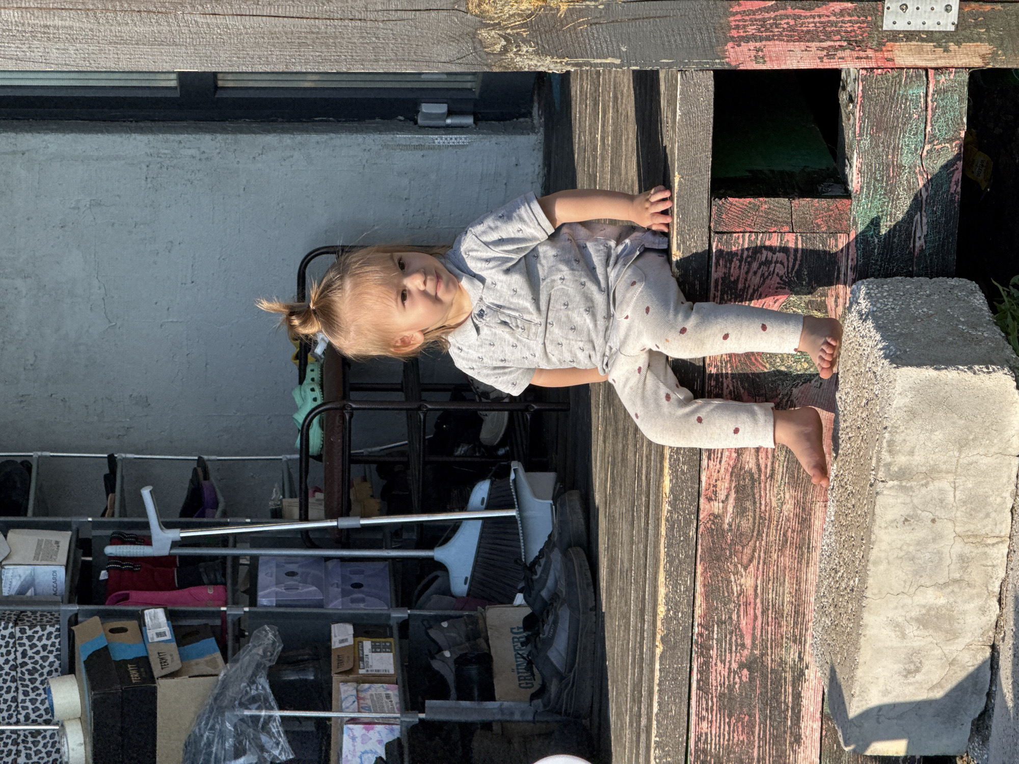 A young child is lying on a wooden surface, possibly a porch or deck. The child is wearing light-colored clothes with small polka dots. The location is potentially Kharnavy, Russia, but this cannot be confirmed. Behind the child, there's a wall with a window and a shelving unit filled with various items, including boxes and cleaning supplies. To the right, there's a concrete block. The wooden surface has been painted and weathered. The child is looking toward the camera.