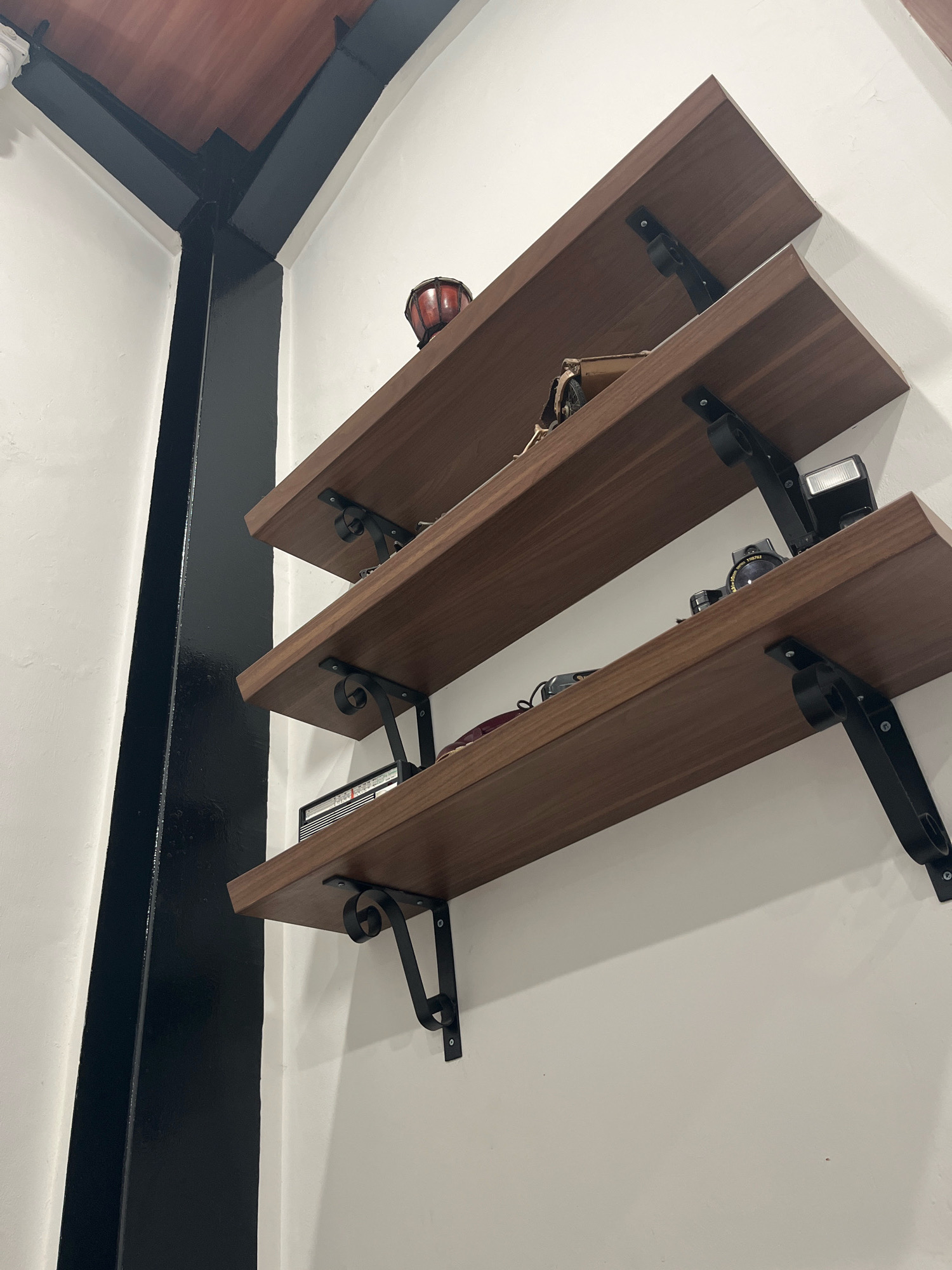 The image shows three wooden shelves mounted on a white wall. The shelves are supported by black metal brackets. On the top shelf, there is a small, red drum-like object and a brown handbag. The middle shelf holds a camera and a camera flash. The bottom shelf has a black and silver radio. A thick black vertical beam is visible on the left side of the image, partially obscuring a wooden ceiling. The scene is indoors, possibly in a home or a shop in Semarang, Indonesia, characterized by its simple and functional display of items.