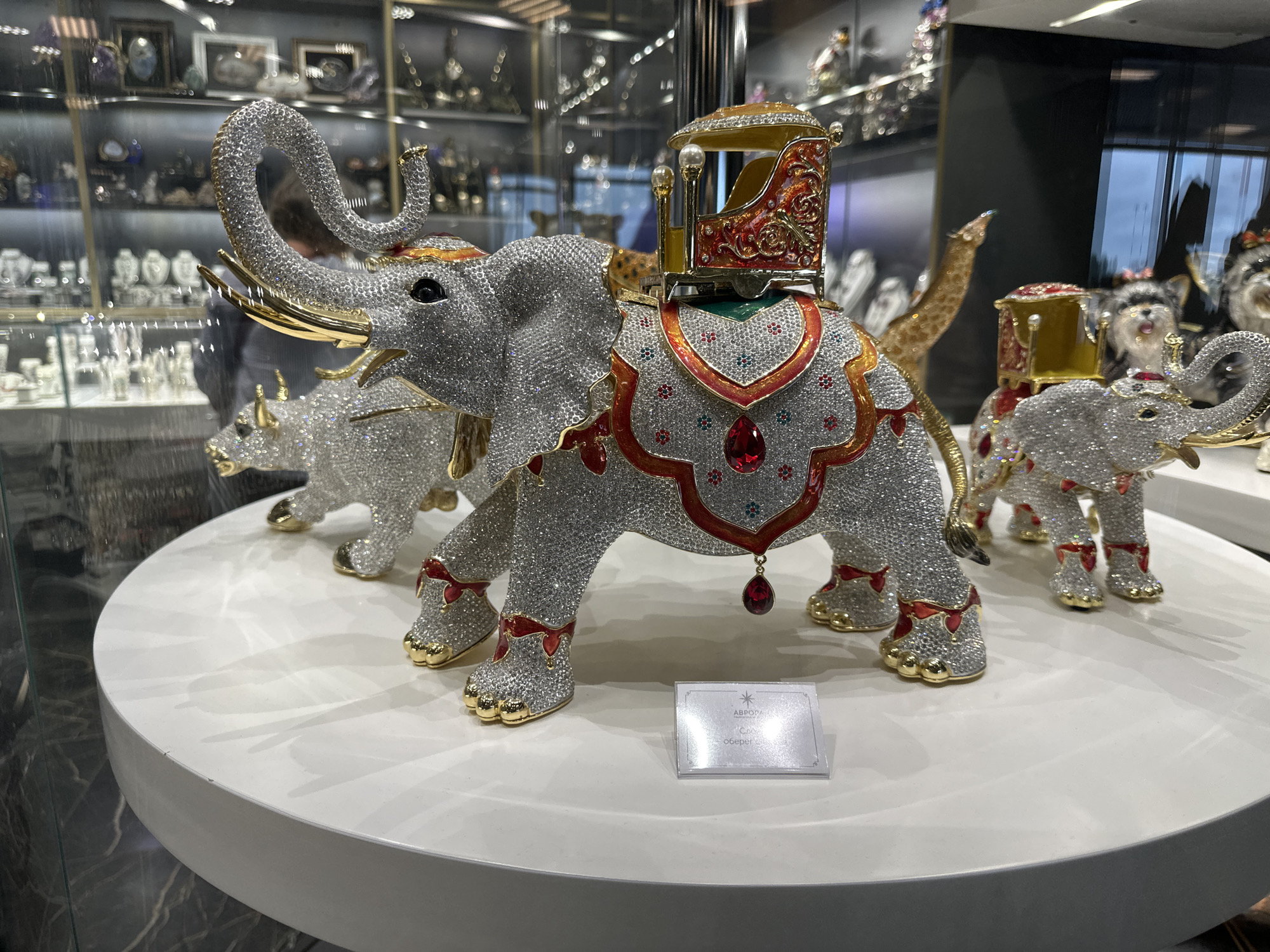 The image depicts a display of bejeweled animal figurines, likely located in a retail setting. The focal point is a large, silver elephant with a red and gold howdah (a seat or carriage) on its back. The elephant is covered in small, sparkling stones, giving it a dazzling appearance. It has golden tusks and detailed decorations, including a red gem hanging from its belly.
Behind the elephant are other similar figurines, including a rhinoceros and another, smaller elephant. The figurines are placed on a round, white display table. A small, rectangular price tag is situated in front of the large elephant.
The background features a glass display case filled with various items, possibly jewelry or other luxury goods. The reflection in the glass suggests an indoor environment, potentially a high-end store.
While the image does not directly confirm the location in Saint Petersburg, Russia, the store logo on the price tag states "Аврора", which is the Russian word for Aurora. The "Аврора" store is a well-known luxury store located in Saint Petersburg, Russia, which suggests that the image was taken there.