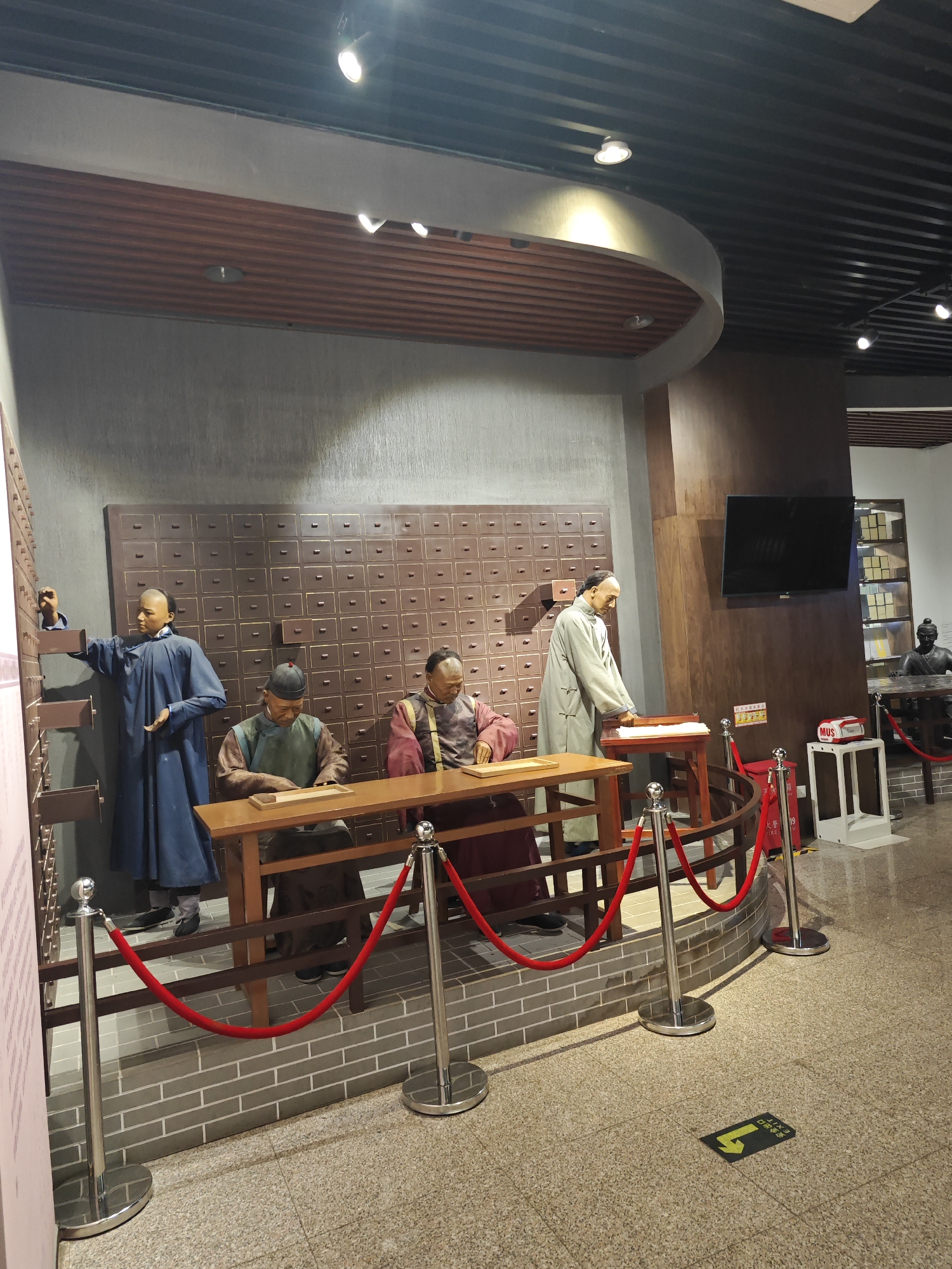 The image depicts a diorama or exhibit likely located in a museum in the Daxing District, China. The exhibit features four mannequin-like figures dressed in traditional Chinese clothing, possibly representing apothecaries or medical practitioners in a historical setting. The figures are positioned in front of a wall with many drawers, suggestive of a traditional Chinese medicine cabinet. There are two tables, one with figures seated and one figure standing to the right of the table. The figures are roped off by silver stanchions with red ropes. The environment includes a textured wall, a television screen mounted on the wall, and an exit sign on the floor. The scene is well-lit and appears to be set in a public display area.