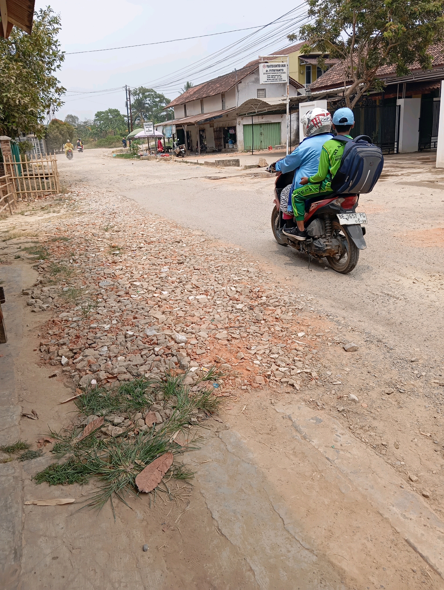 In the city of Leman Benawi, Indonesia, a motorcycle with two occupants travels on a dirt road. The driver, wearing a blue shirt and helmet, is accompanied by a passenger in a green shirt and blue cap, who has a backpack on their back. The road is unpaved and appears to be under construction or repair, with scattered gravel and debris. Buildings line the sides of the road, some with open storefronts. Vegetation, including trees and grass, is visible throughout the scene. The sky is overcast.