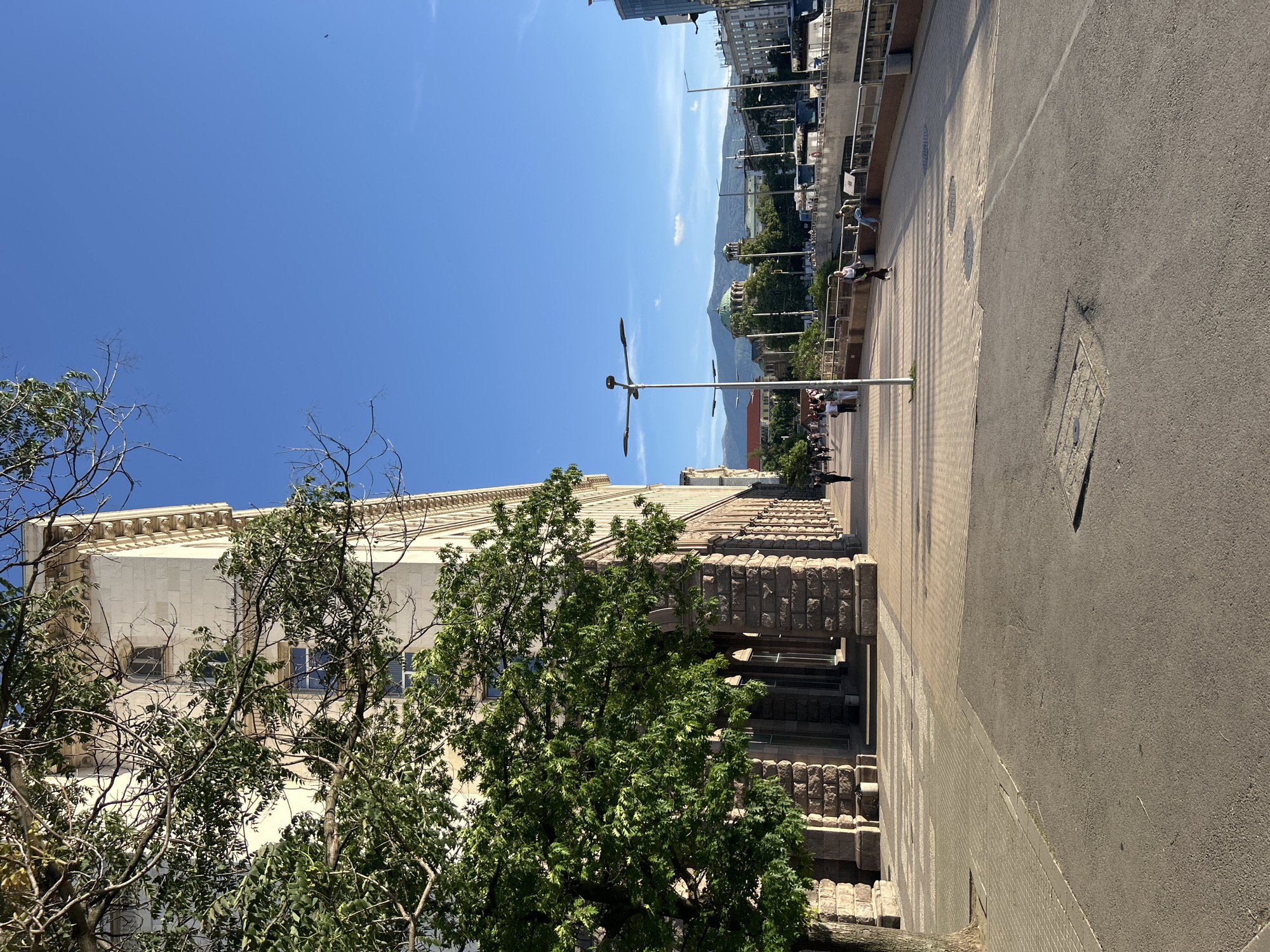 The image captures a city street scene on a clear, sunny day. A large building occupies the left side of the frame, partially obscured by trees. Its facade features light-colored stone with decorative architectural elements. To the right of the building, a wide pedestrian walkway is visible, transitioning into a paved street. Several people are walking along the walkway. In the distance, a mountain range rises above the cityscape, punctuated by a prominent green dome. Streetlights line the walkway. The sky is a clear blue.