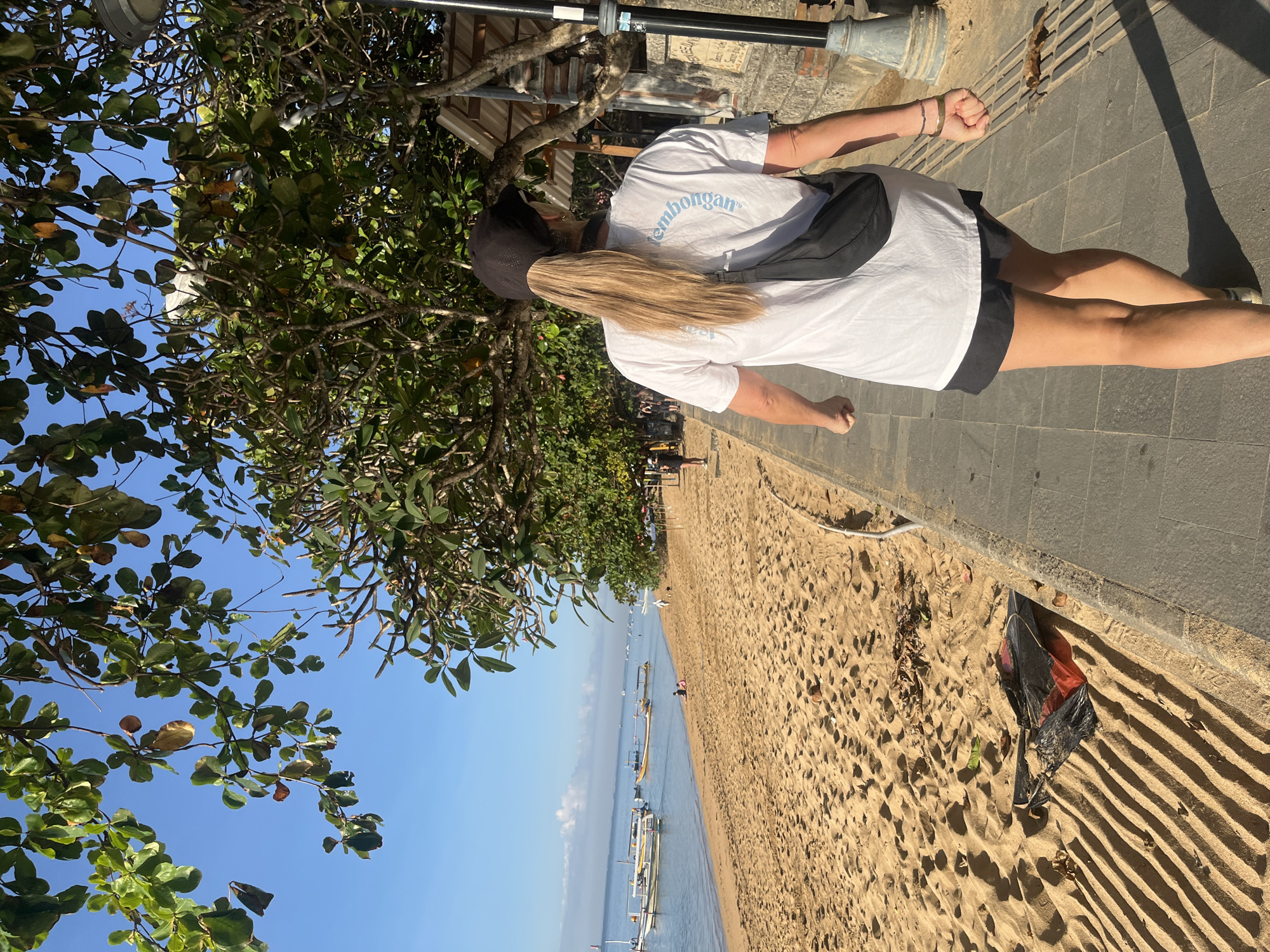 The image depicts a sunlit beach scene in Denpasar, Indonesia. The foreground features a woman with blonde hair in a ponytail, wearing a black baseball cap, a white t-shirt with blue text on the back, and black shorts. She is walking away from the camera on a paved walkway that borders a sandy beach. A black fanny pack is slung across her chest. The beach itself is golden sand, with some debris, including a crumpled black and red object, lying on it. In the background, the calm blue ocean is dotted with several traditional Balinese boats. Beyond the beach, lush green trees with dappled sunlight filter through their leaves, and a small building with a thatched roof is visible. The sky is clear and blue.