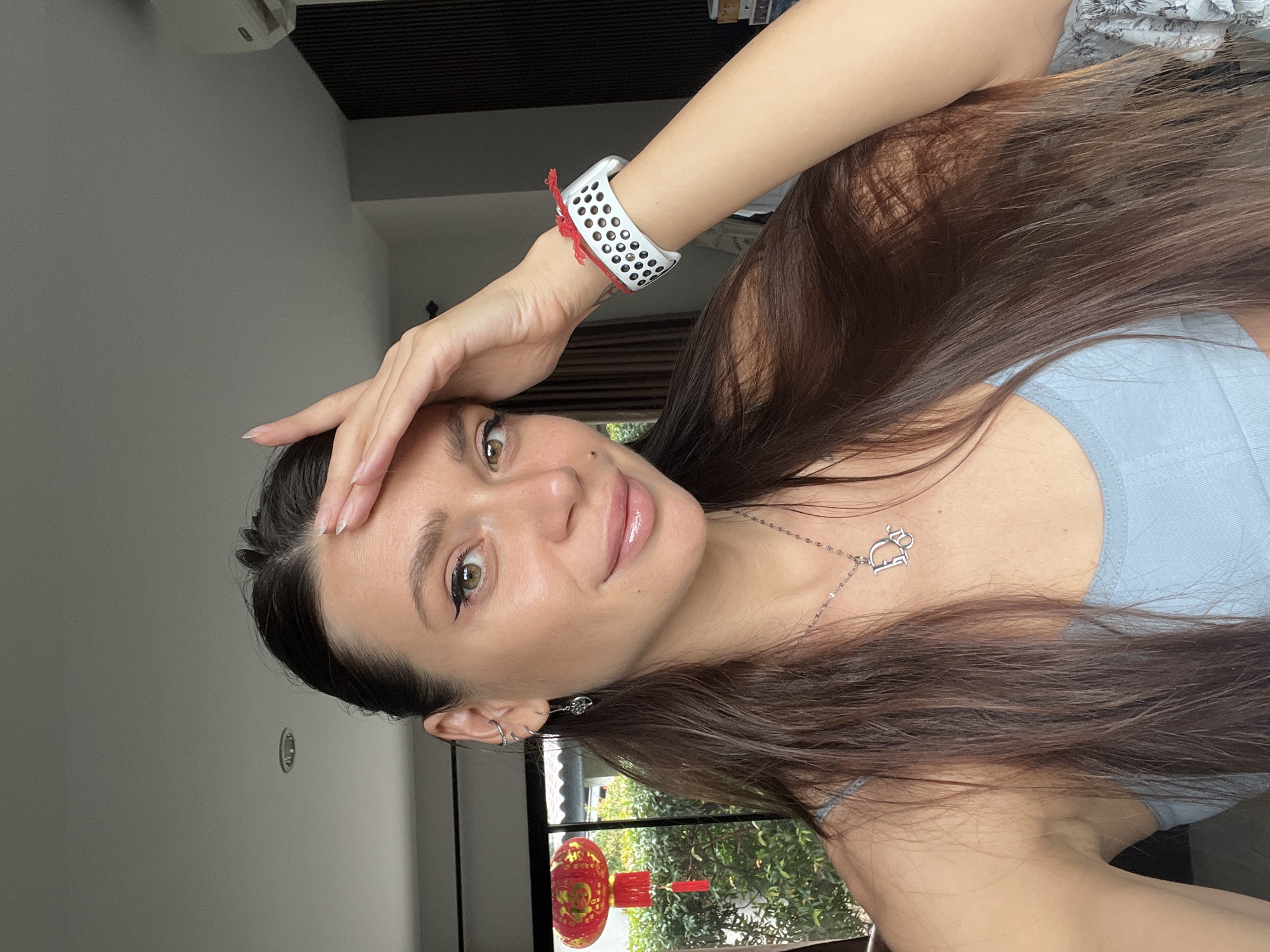 A woman is seen from the chest up, with her right hand touching her forehead. She has long brown hair, a light blue top, and a silver necklace with a letter "D" pendant. She is wearing a white Apple Watch with a red string tied to it. Outside the window behind her, there is a red Chinese lantern hanging, suggesting a location in a city with a Chinese community or influence. The exact location is unknown.